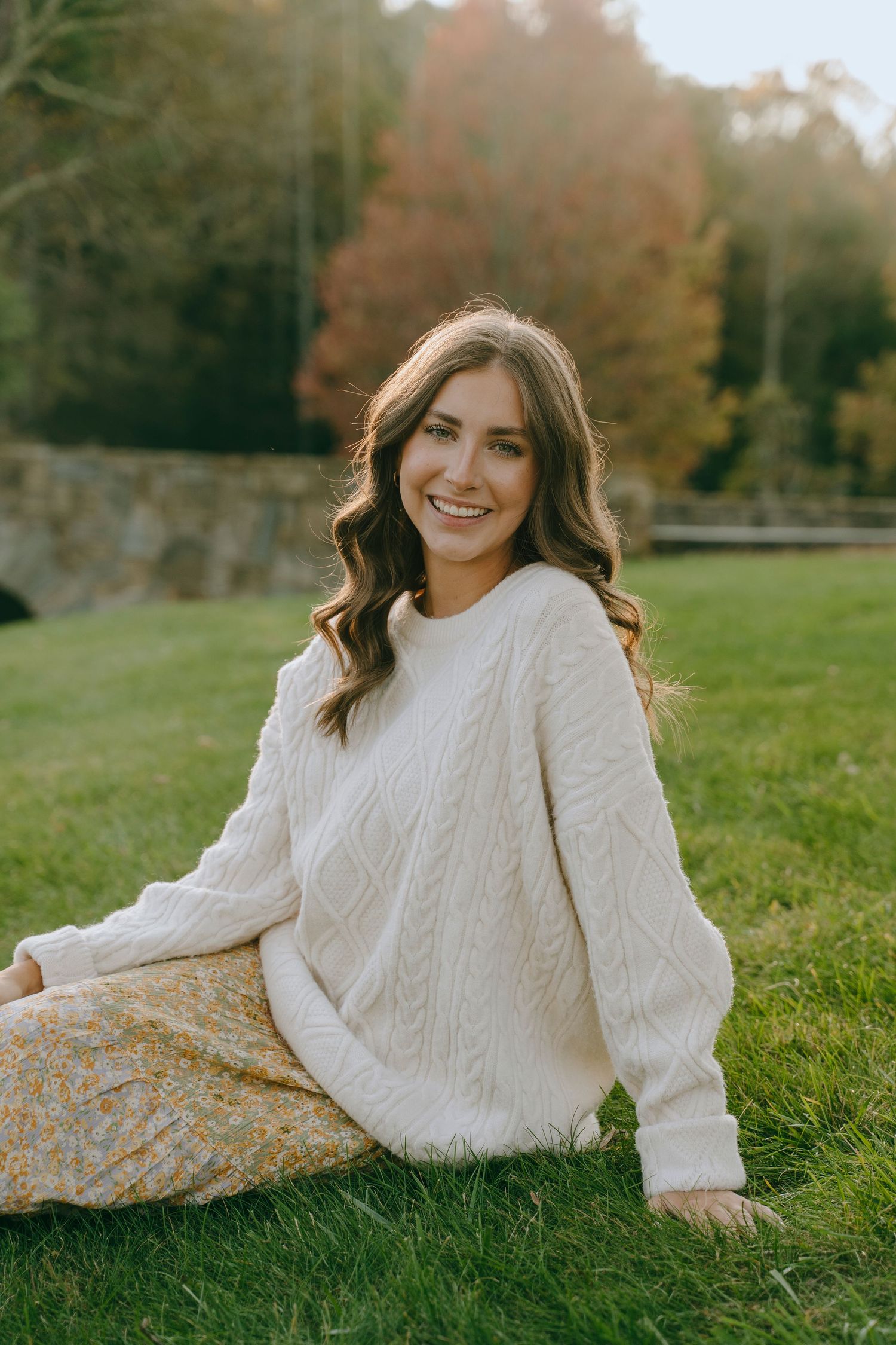 Outdoor lifestyle portrait of a smiling woman in a white sweater and floral skirt, sitting on the grass in warm sunlight.