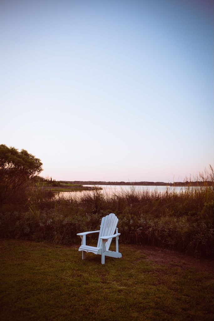 A white Adirondack chair sits alone on a grassy hill overlooking a serene lake at sunset with bushes in view.