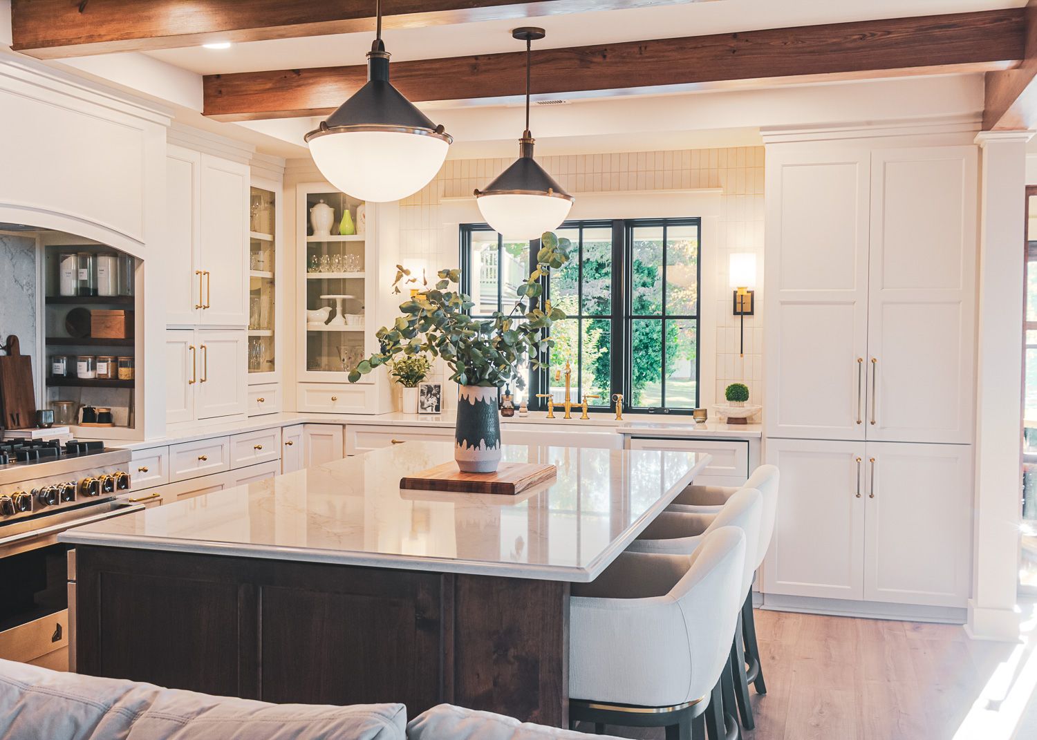 Modern white kitchen with wood beams, black countertops, pendant lighting, and large windows offering natural light.