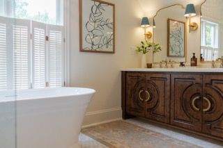 Elegant master bathroom with white freestanding tub, dark wood vanity, and natural light streaming through shuttered windows.