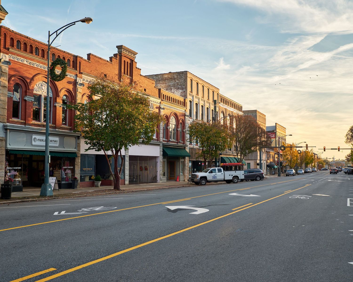 Historic brick buildings line both sides of a wide main street in a small downtown business district at sunset.