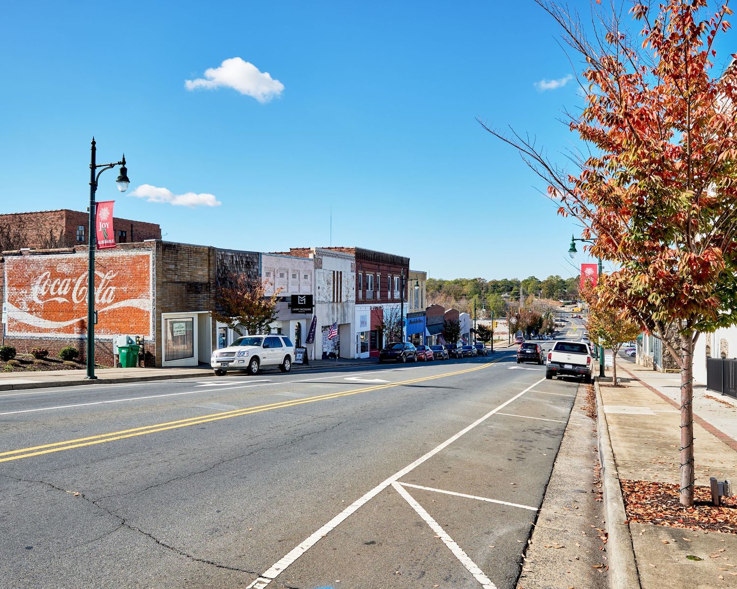 Small town main street with brick buildings, autumn trees, and vintage Coca-Cola sign on a sunny day.