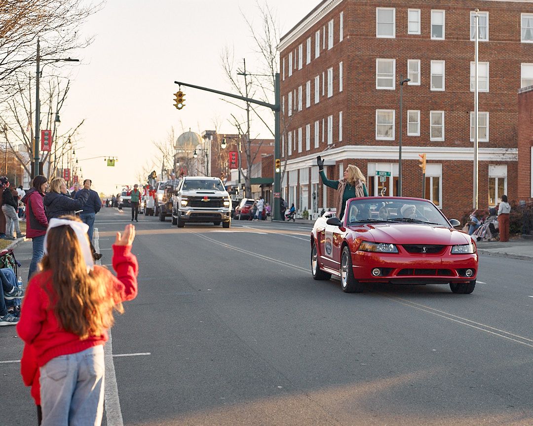 Spectators watch a red sports car drive through a small town intersection during a parade or event.