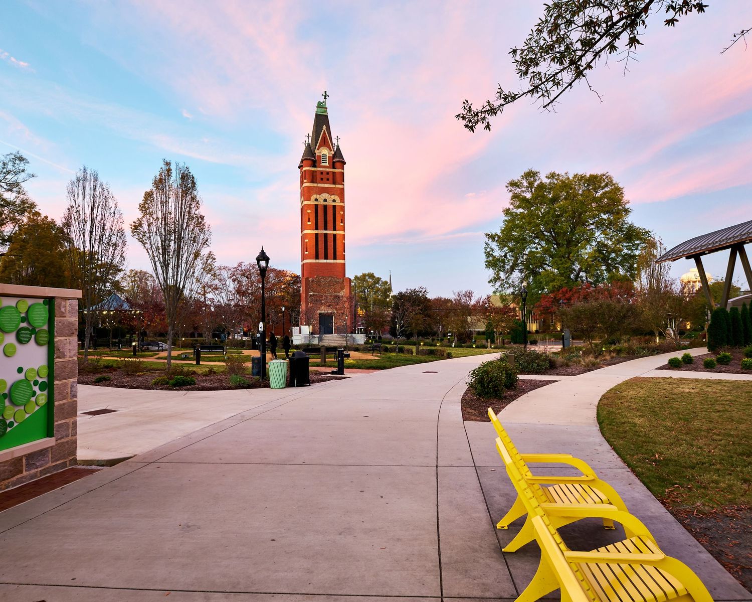 A panoramic view of a college campus with a clock tower and yellow bench under pink sunset skies.