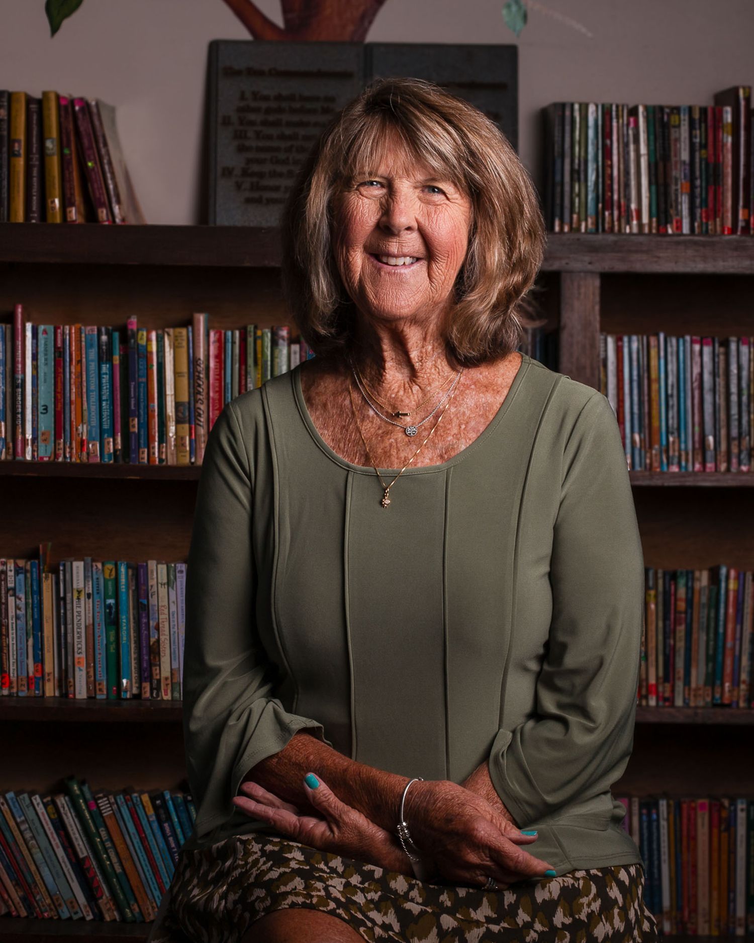 A woman in an olive green top sits in front of bookshelves in a library setting with dramatic lighting.