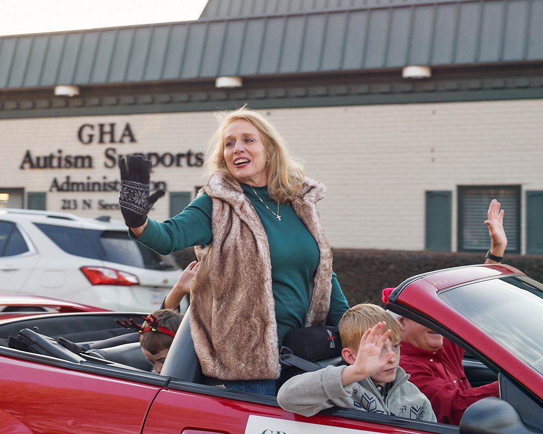 A group of people wave and smile from inside vehicles during what appears to be a drive-through celebration event.