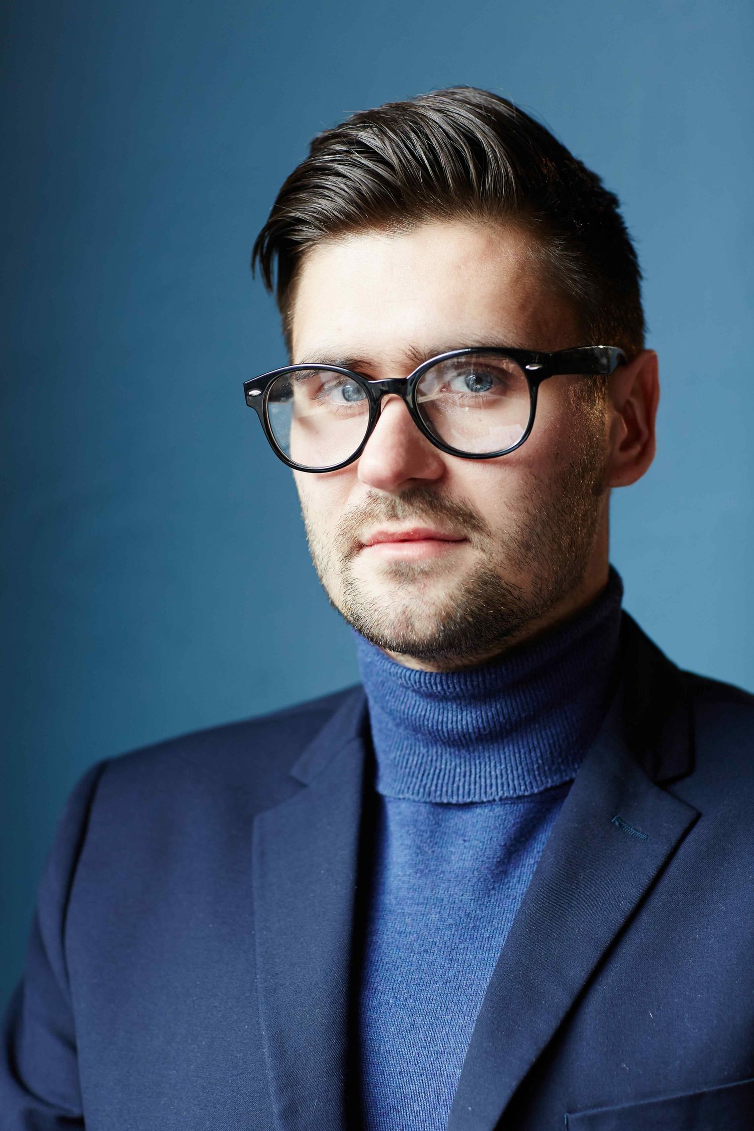 Professional studio portrait of someone wearing glasses, a navy blazer and blue turtleneck sweater against blue backdrop.