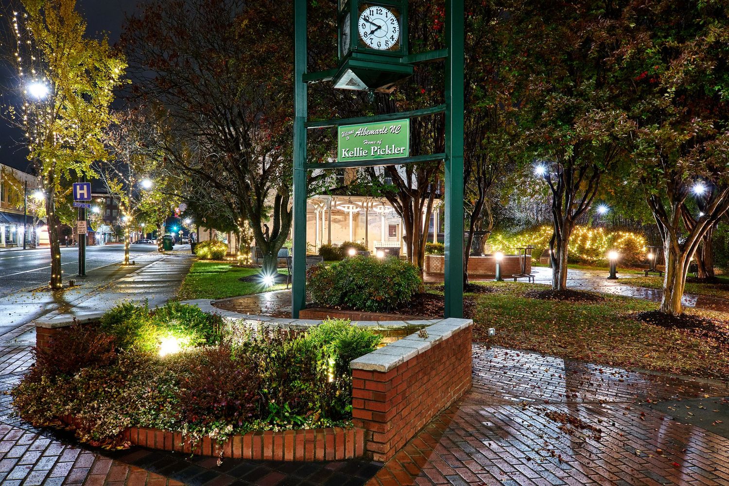 A scenic nighttime view of illuminated brick pathways winding through a lush garden with lampposts and trees.