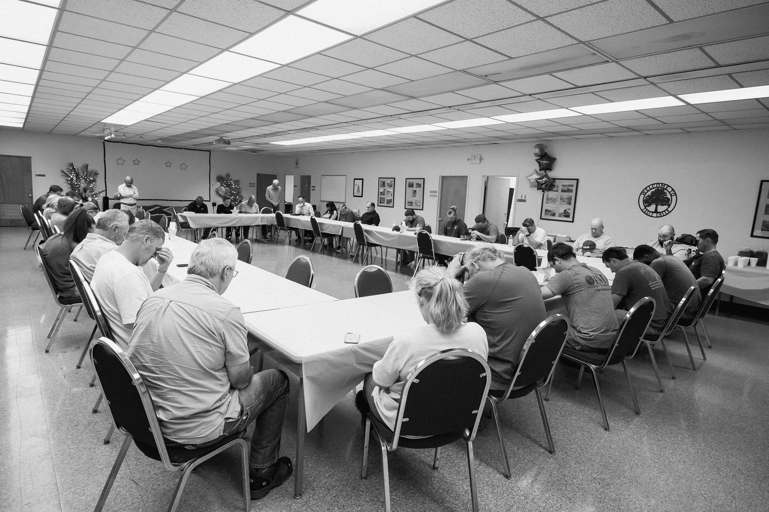 People seated at tables in a classroom or meeting room with fluorescent lighting and white ceiling tiles.