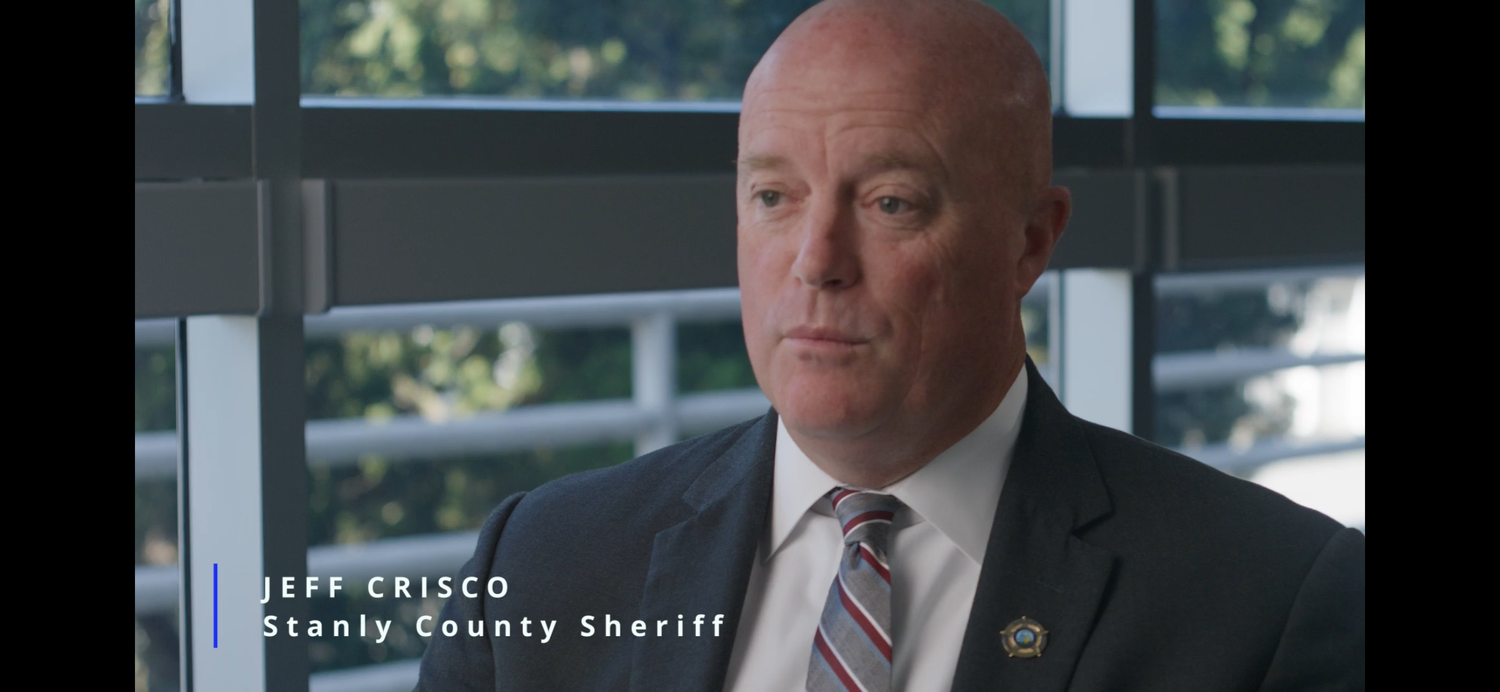 Law enforcement official seen speaking in professional indoor setting with natural light coming through windows.