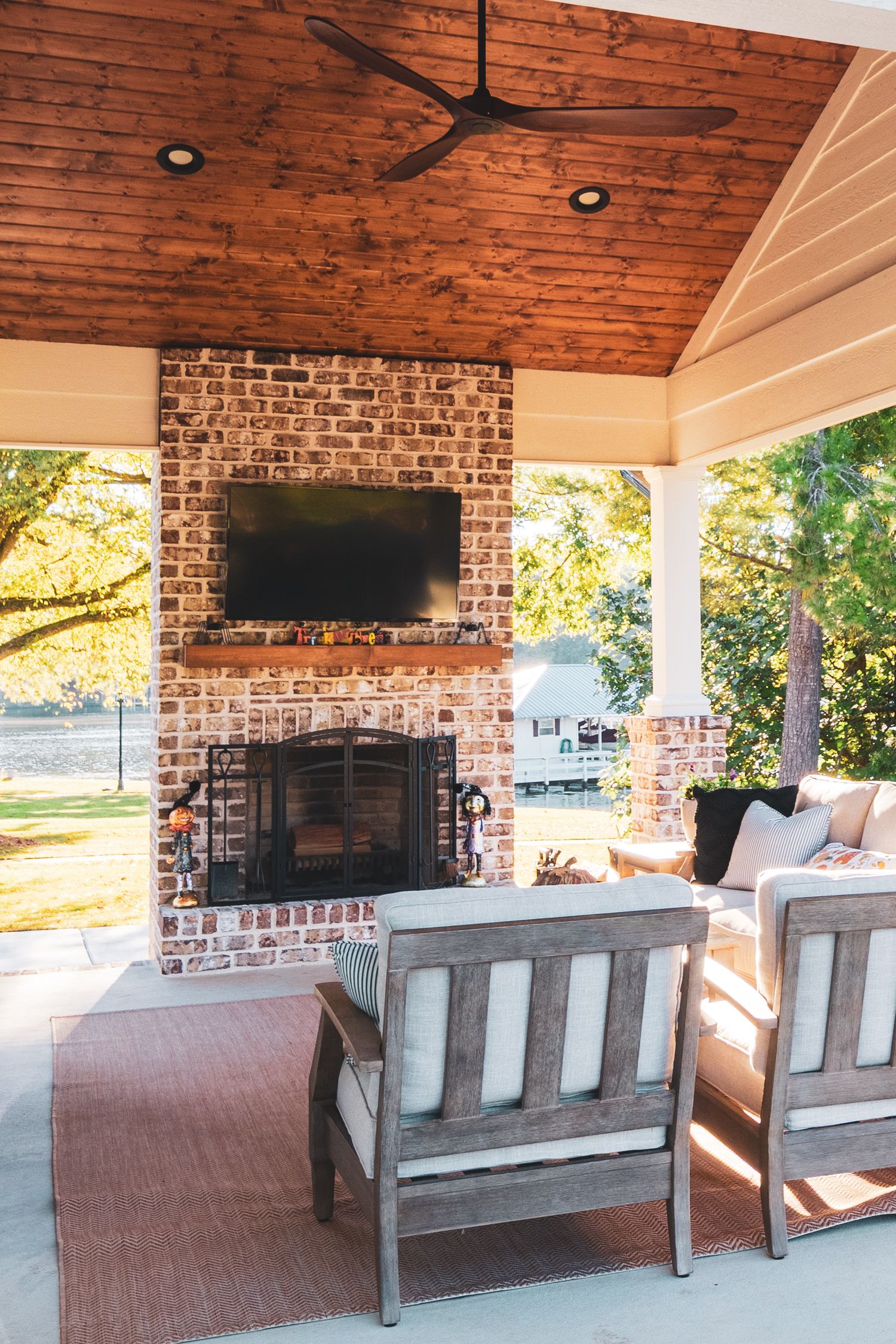 A covered outdoor patio with wooden ceiling, brick walls, mounted TV, and gray patio furniture creates a cozy living space.