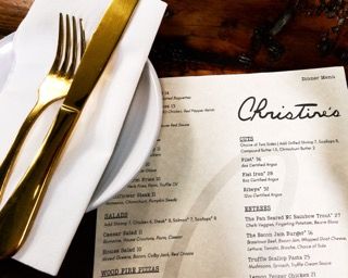 Restaurant menu from Christine's with gold cutlery and white plate on dark wooden table.