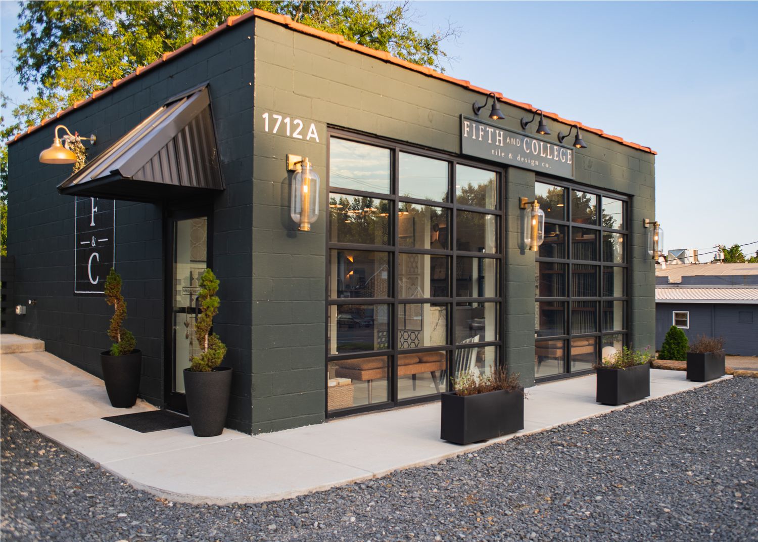 Modern small commercial building with dark gray exterior, large windows, and black awning featuring potted plants and signage.
