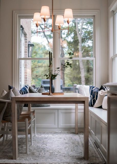 Cozy breakfast nook with built-in bench seating, wooden table, and elegant chandelier by large windows overlooking trees.