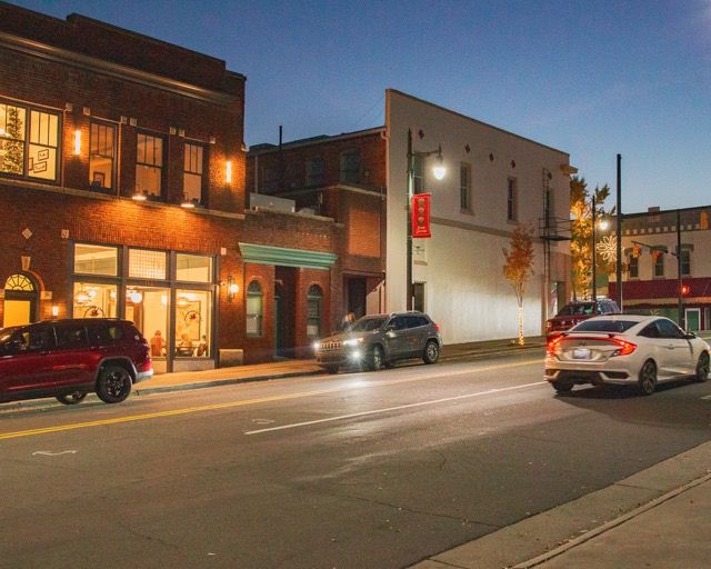 Small town main street at dusk with illuminated storefronts and parked cars along a quiet commercial district.