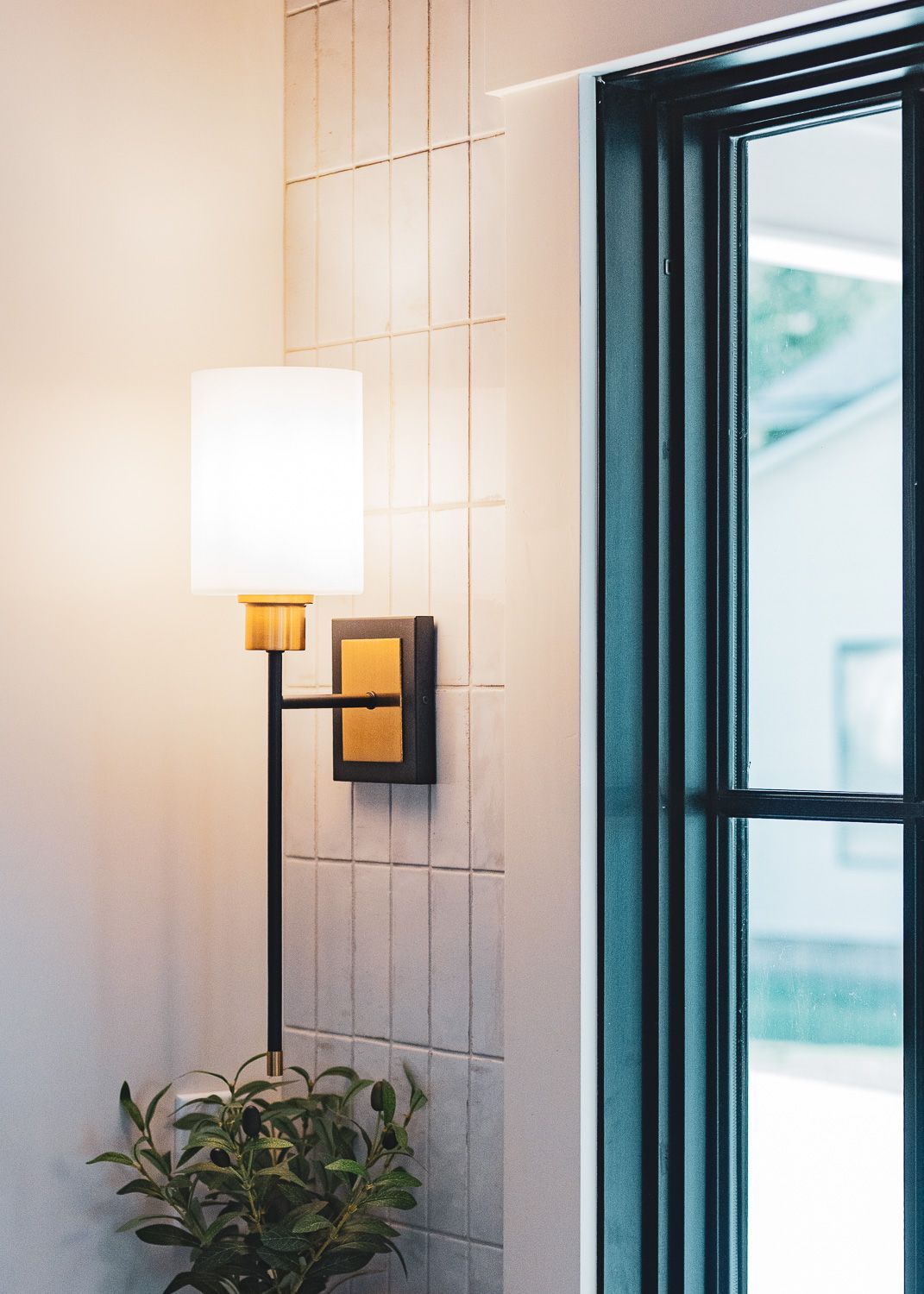 Modern wall sconce lighting fixture mounted on a white textured wall with green window frames visible.