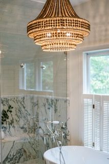 A modern bathroom features a crystal chandelier, marble walls, and white window shutters creating an elegant atmosphere.