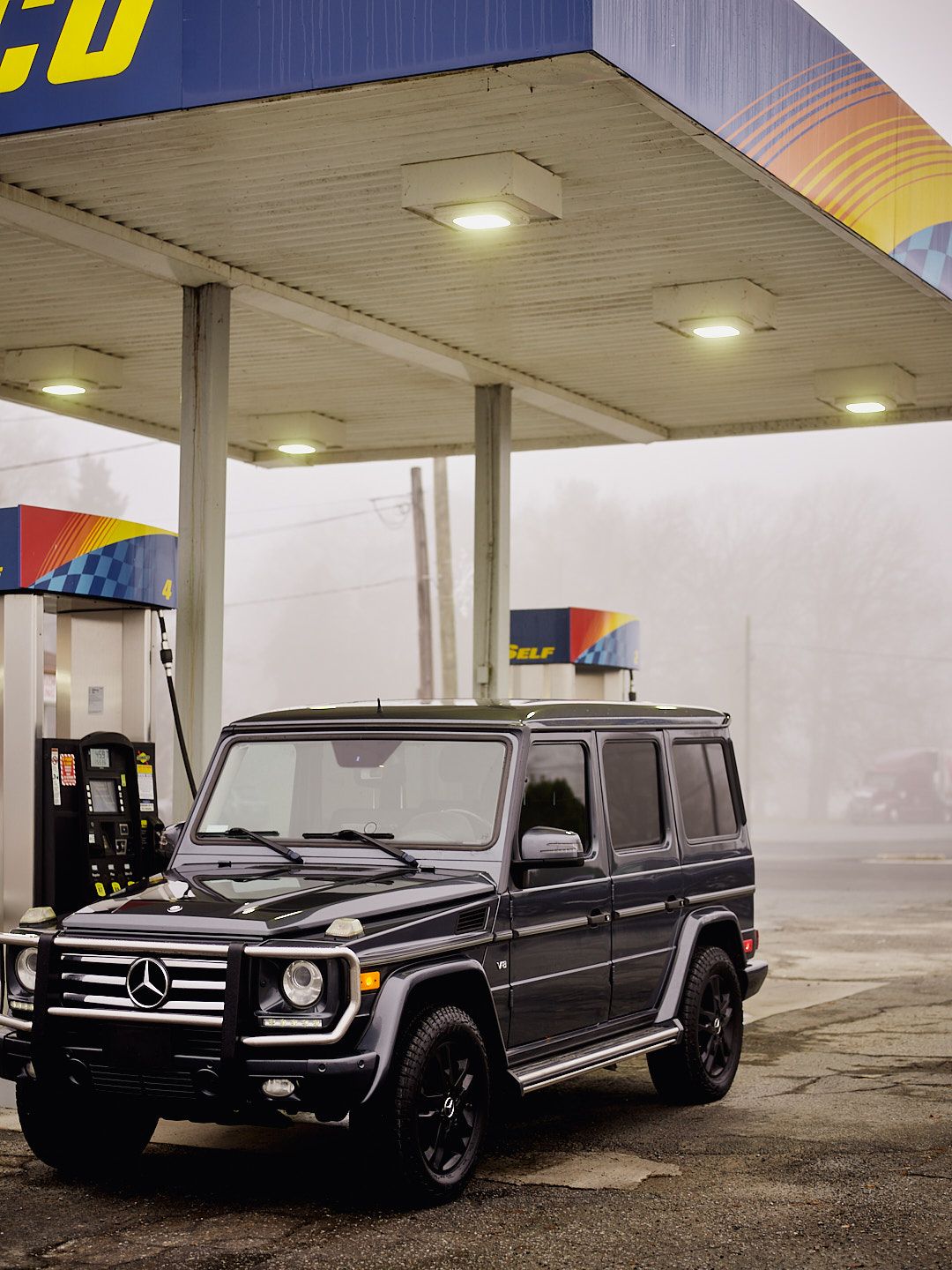 Black Mercedes-Benz G-Class SUV parked at a gas station in foggy conditions with illuminated overhead lights.