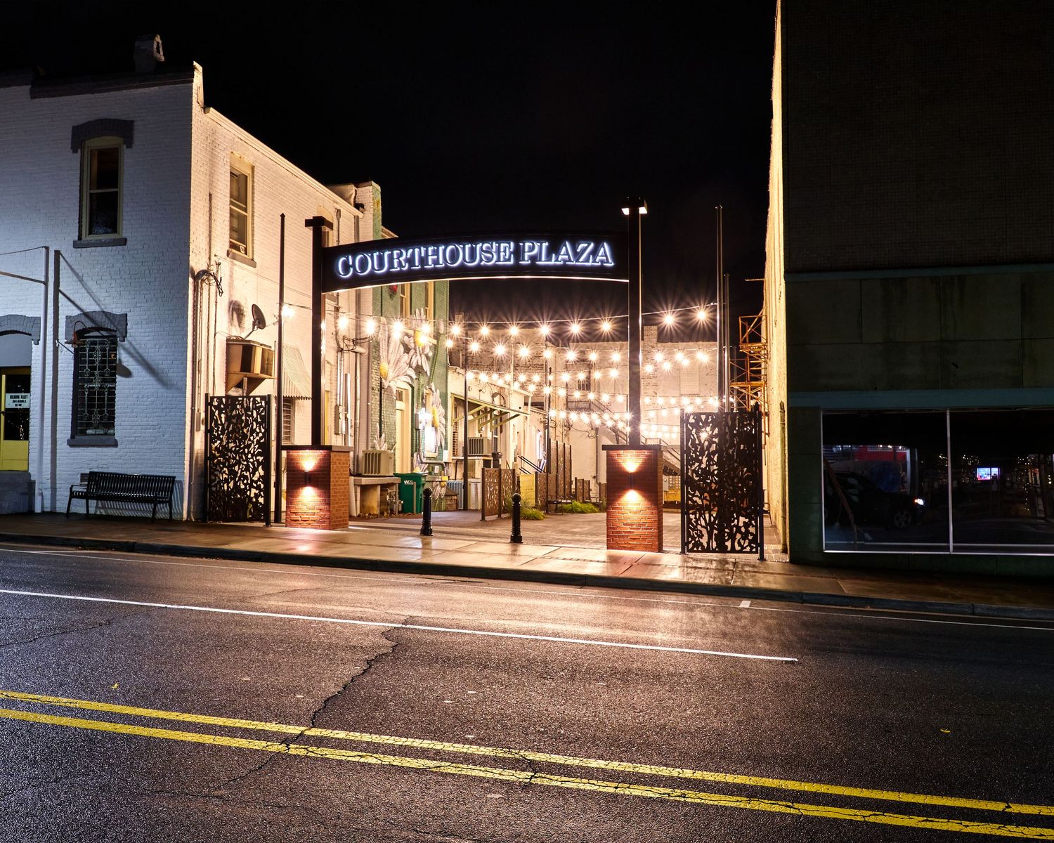 Illuminated entrance to Sportsmens Plaza at night with string lights and glowing signage along a city street.