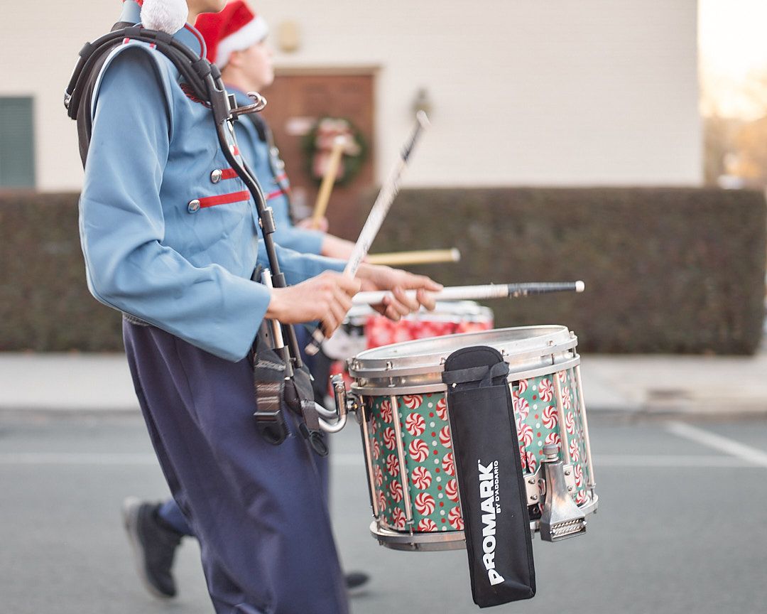 Marching band drummer wearing light blue uniform and navy pants plays snare drum in parking lot during practice.