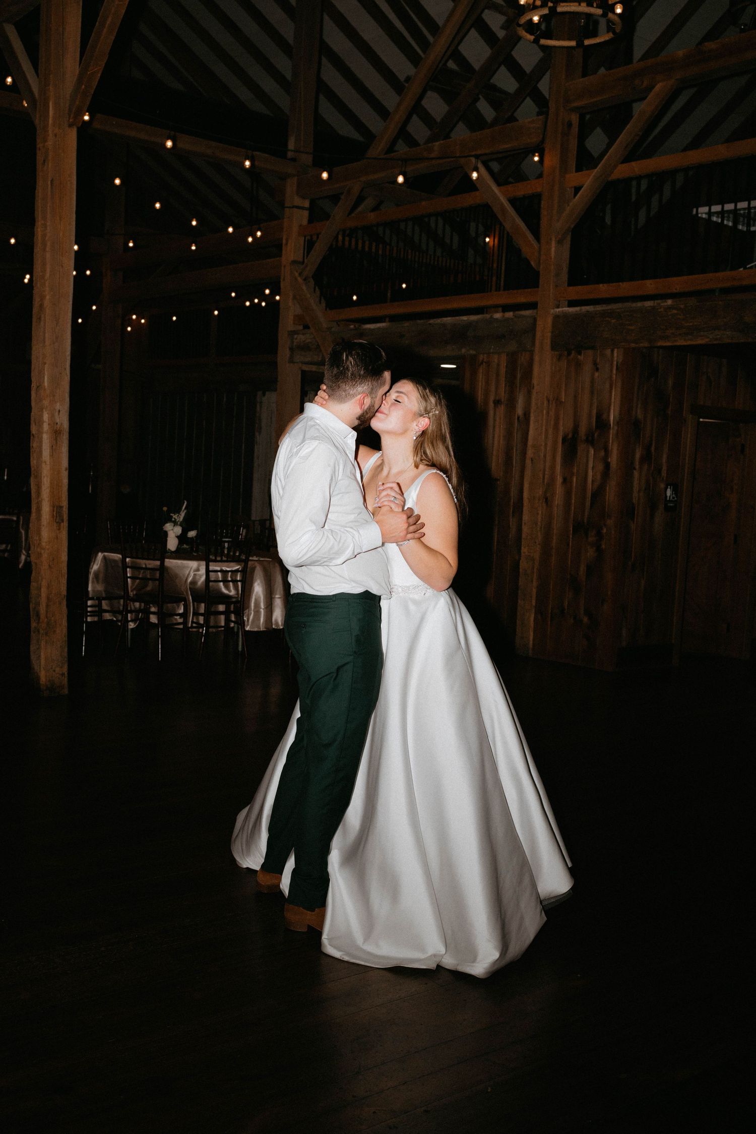 Couple shares a romantic first dance in a dimly lit rustic barn with twinkling lights above.