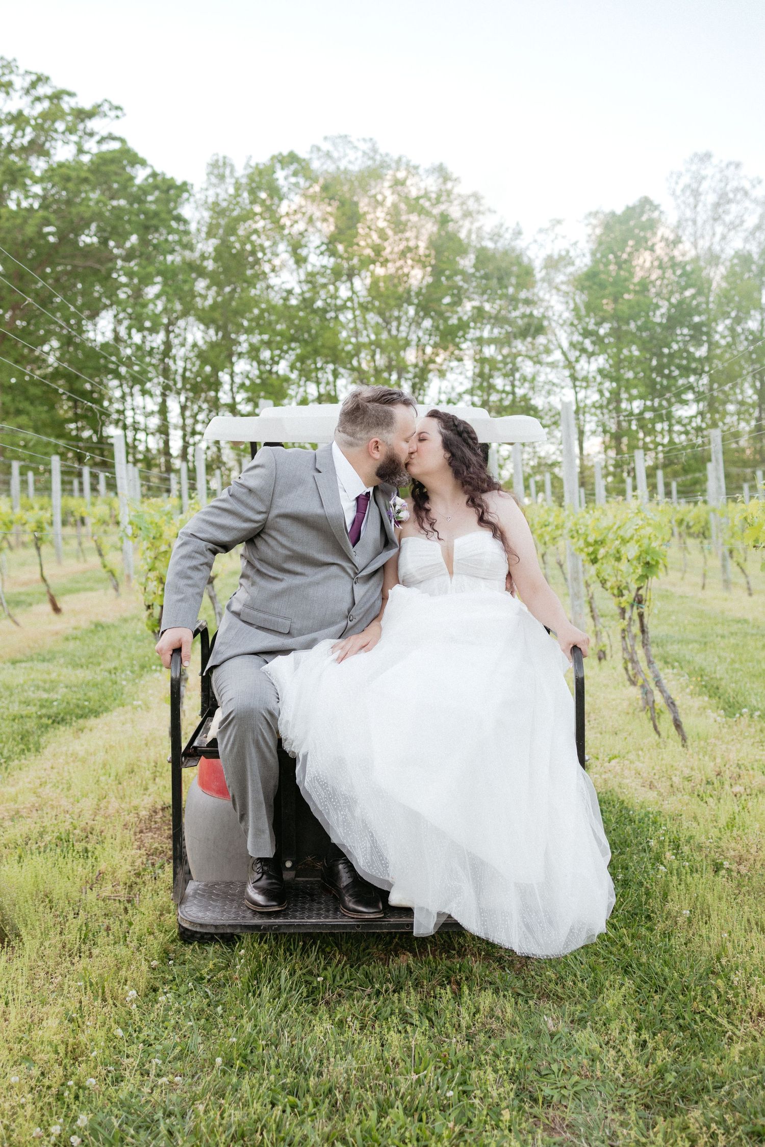 A bride and groom share a romantic kiss while sitting in a vintage car in a vineyard setting.