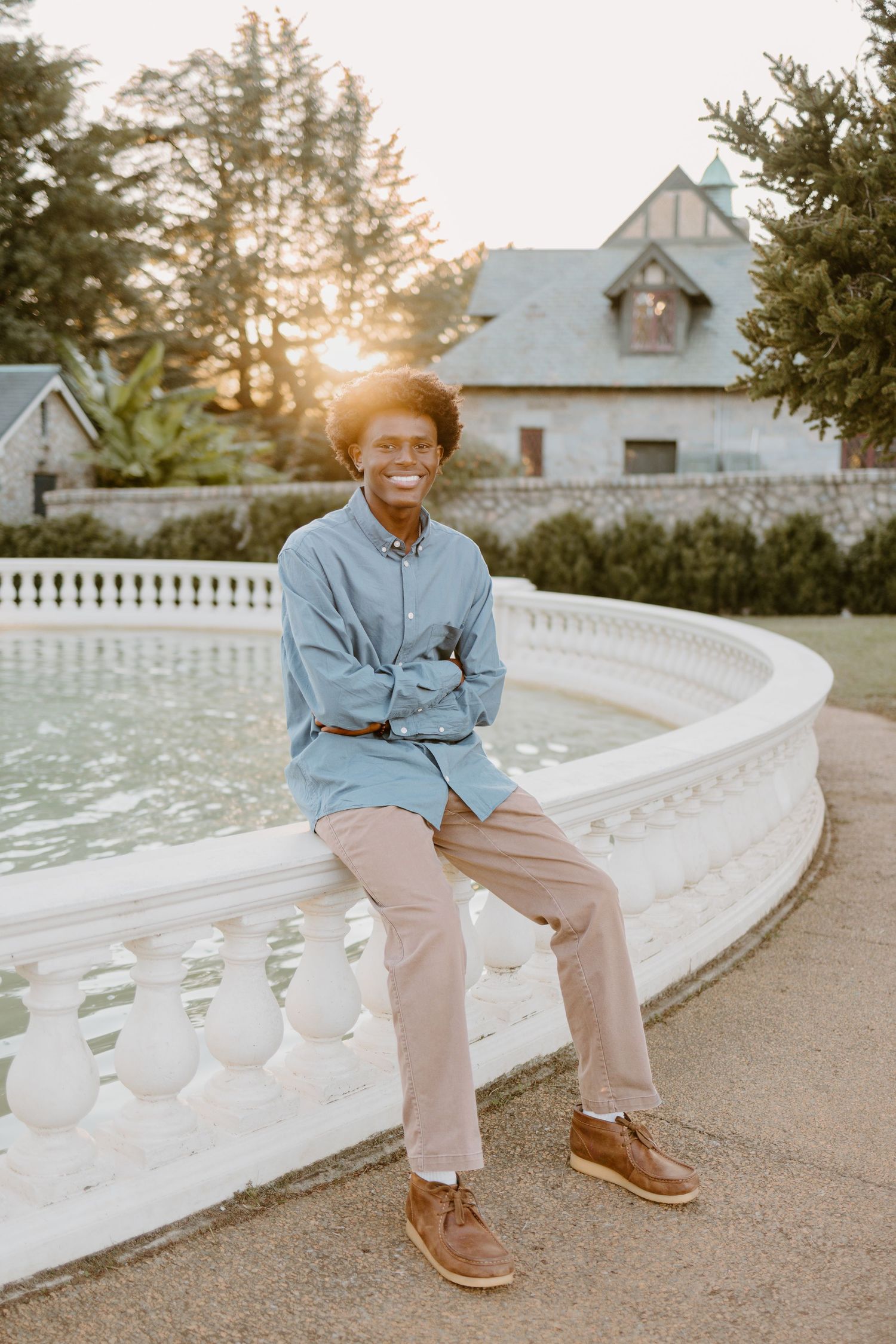 Person in light blue jacket and khaki pants sits on curved white balustrade at sunset.