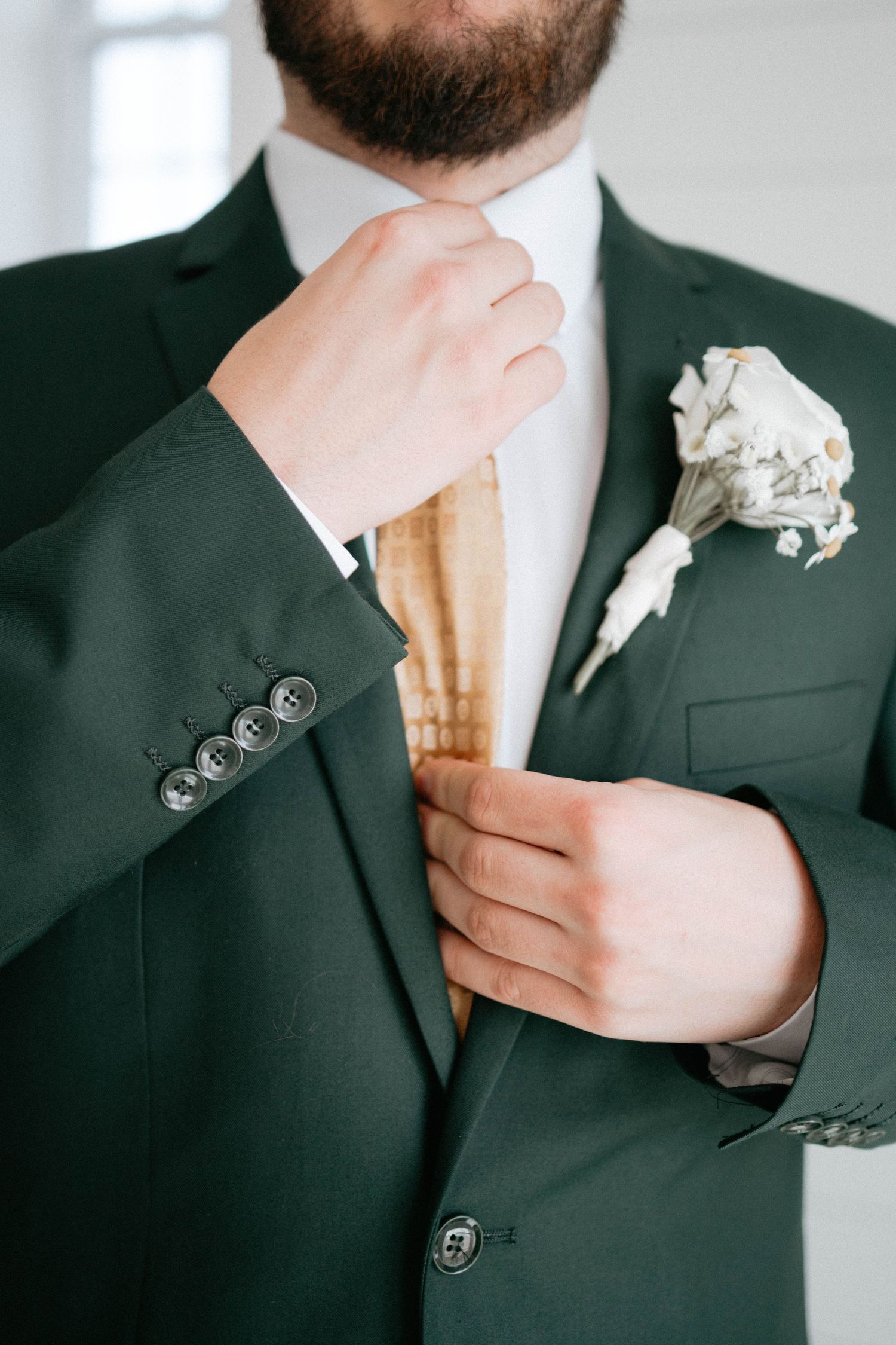 Close-up of groom in black suit adjusting his tie while showing white boutonniere flower detail.
