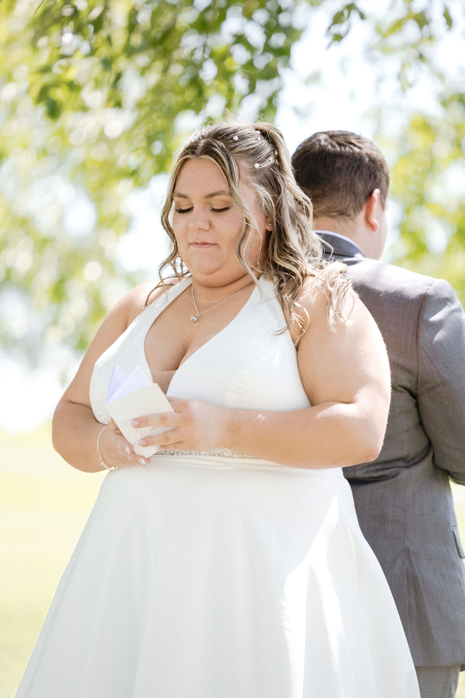 A romantic wedding couple embraces outdoors in a sun-dappled setting with natural greenery and trees.
