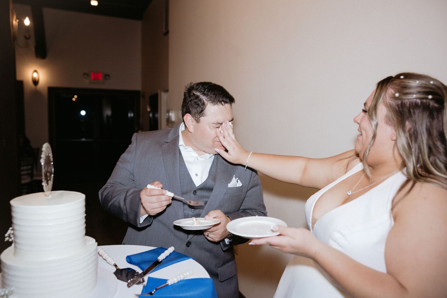 A playful cake feeding moment at a wedding reception with a white tiered cake in the foreground.