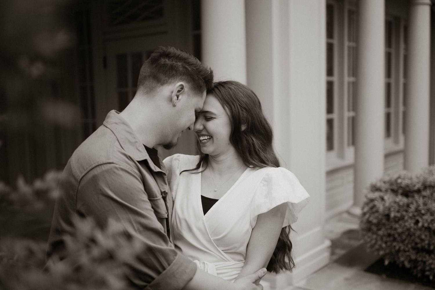 Romantic black and white portrait of a couple sharing an intimate moment outdoors by a pillared building entrance.