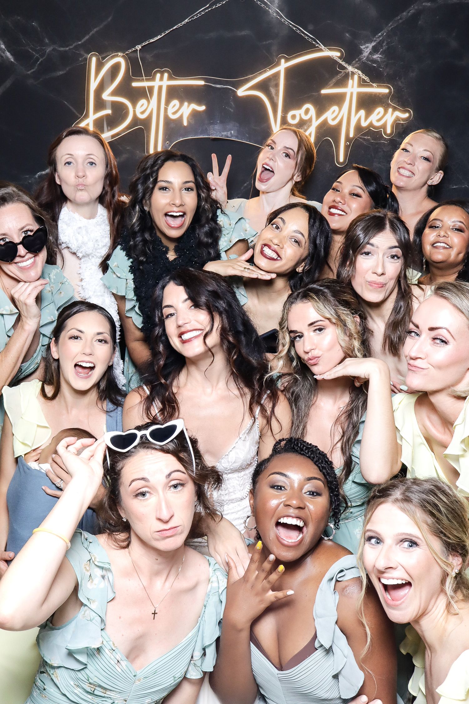 Large group of bridesmaids in pastel dresses posing playfully under a neon sign at a wedding celebration.