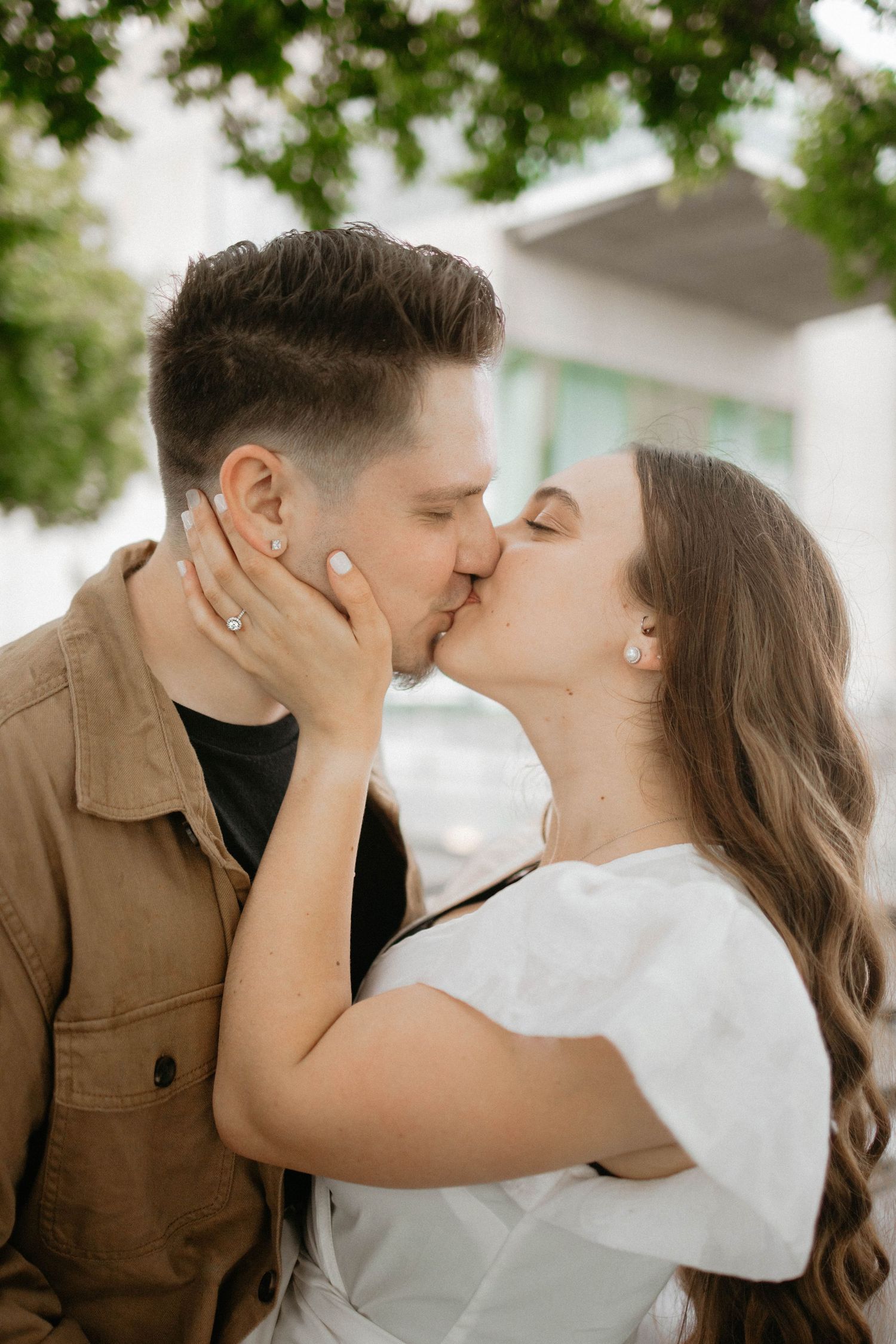 A romantic couple sharing tender kisses while embracing outdoors under green foliage.