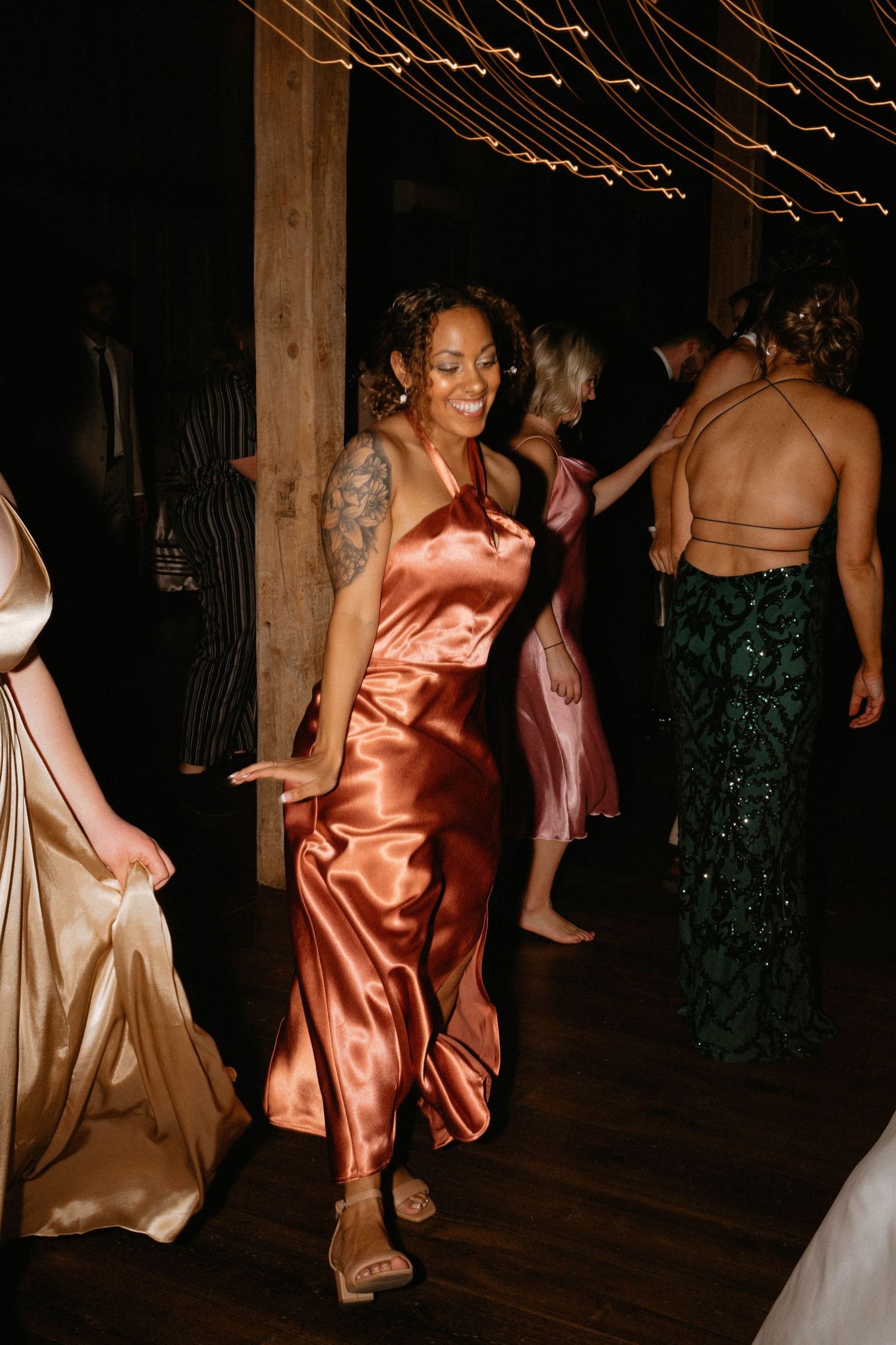 Wedding reception dance party with guests in satin dresses dancing under dim lighting and string lights.