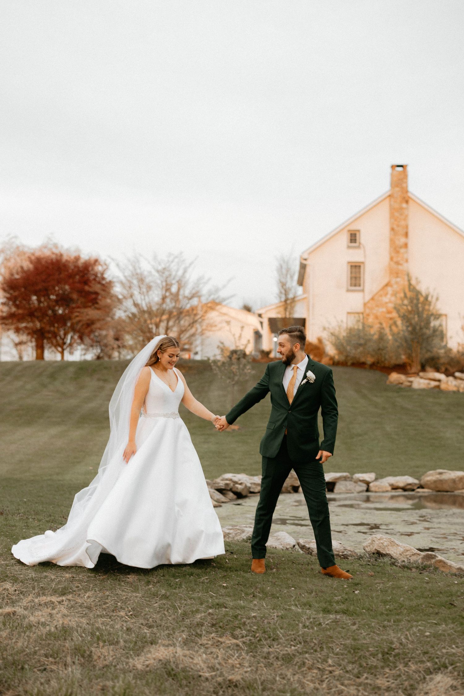 Wedding couple playfully interacts on grassy hill near rustic stone farmhouse.