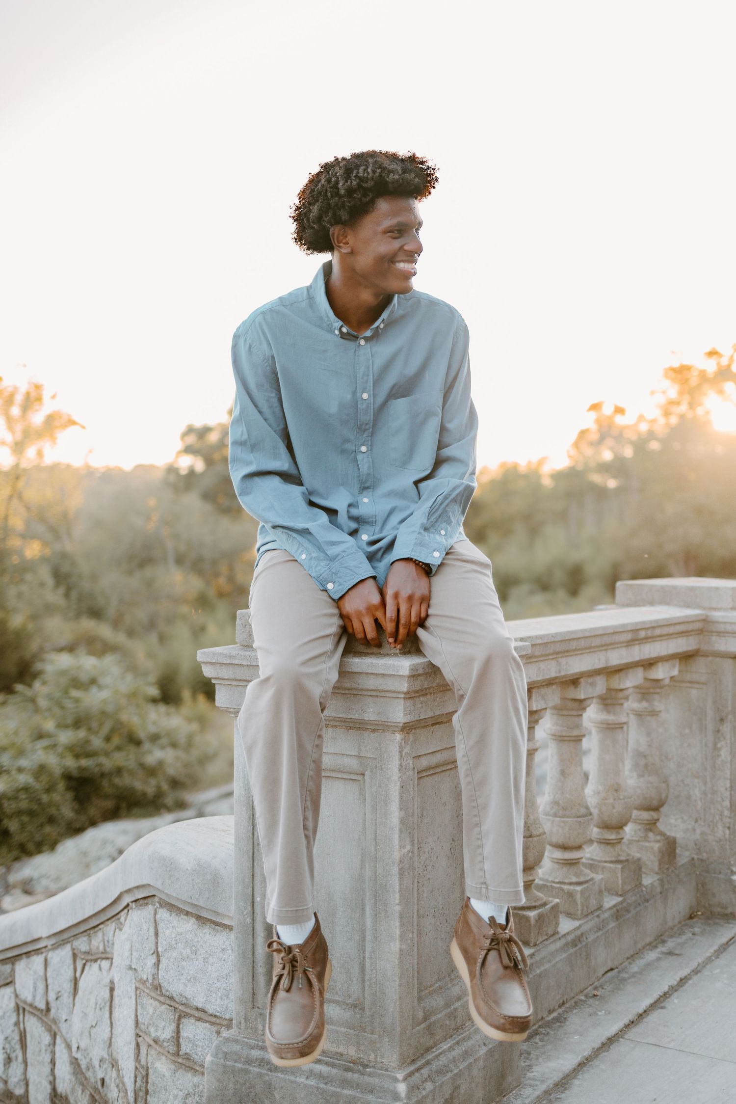 Person in light blue shirt and white pants sits casually on stone balustrade during golden sunset.