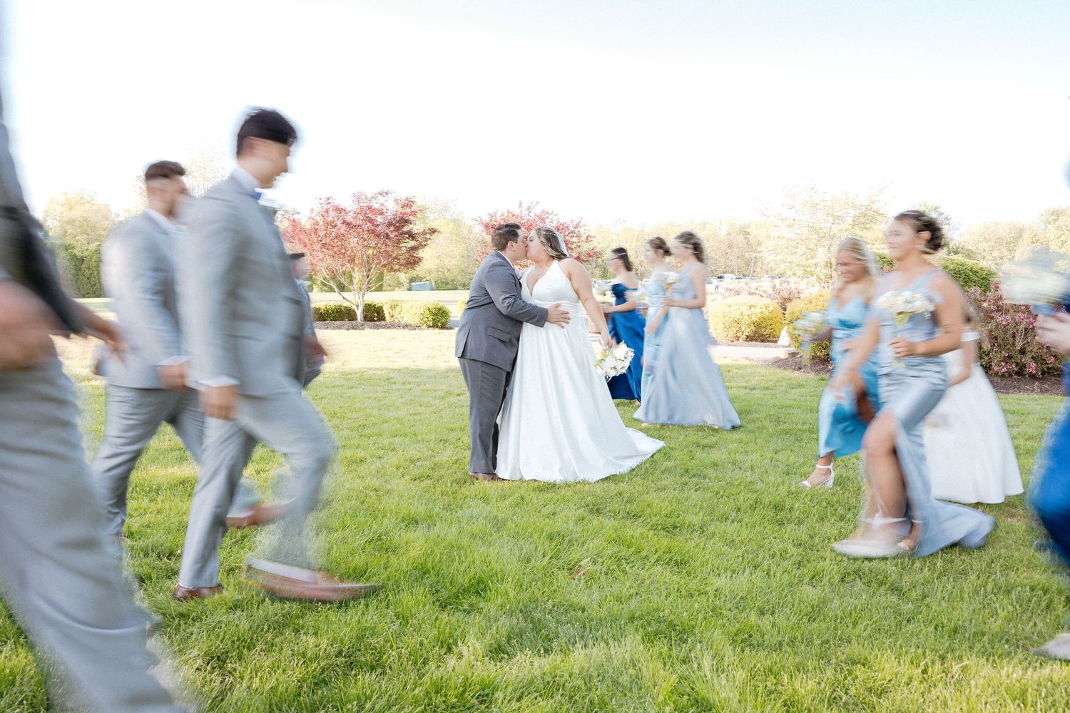 Motion-blurred wedding party dancing on grass with guests in blue and grey attire.