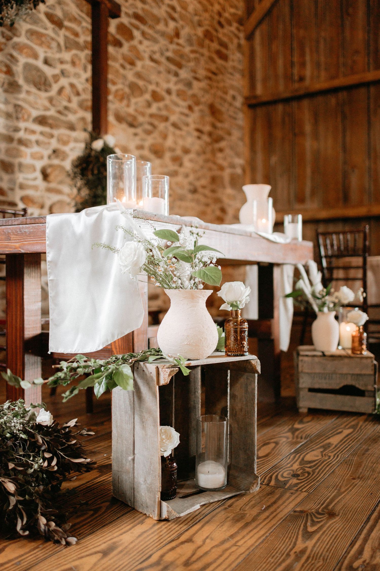 White floral centerpieces and vases arranged on wooden tables in a stone-walled rustic venue.