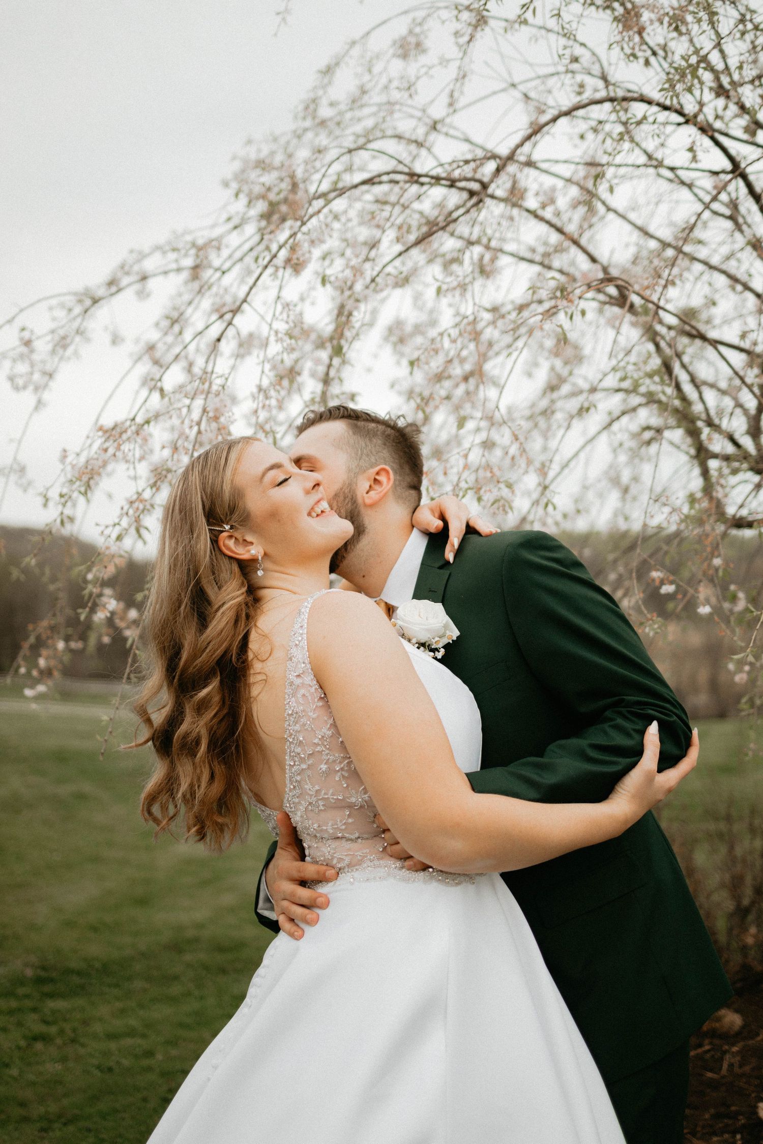 A couple in wedding attire embrace and kiss under flowering cherry blossom trees in a romantic springtime setting.