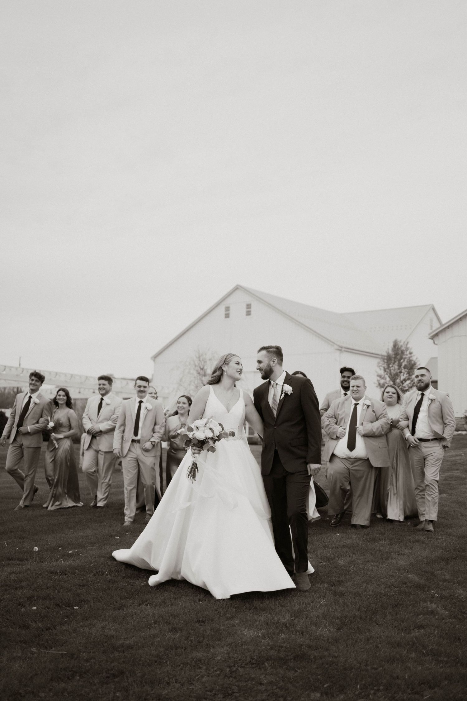 A wedding party runs playfully across a grassy field with a white tent in the background, creating a joyful outdoor scene.