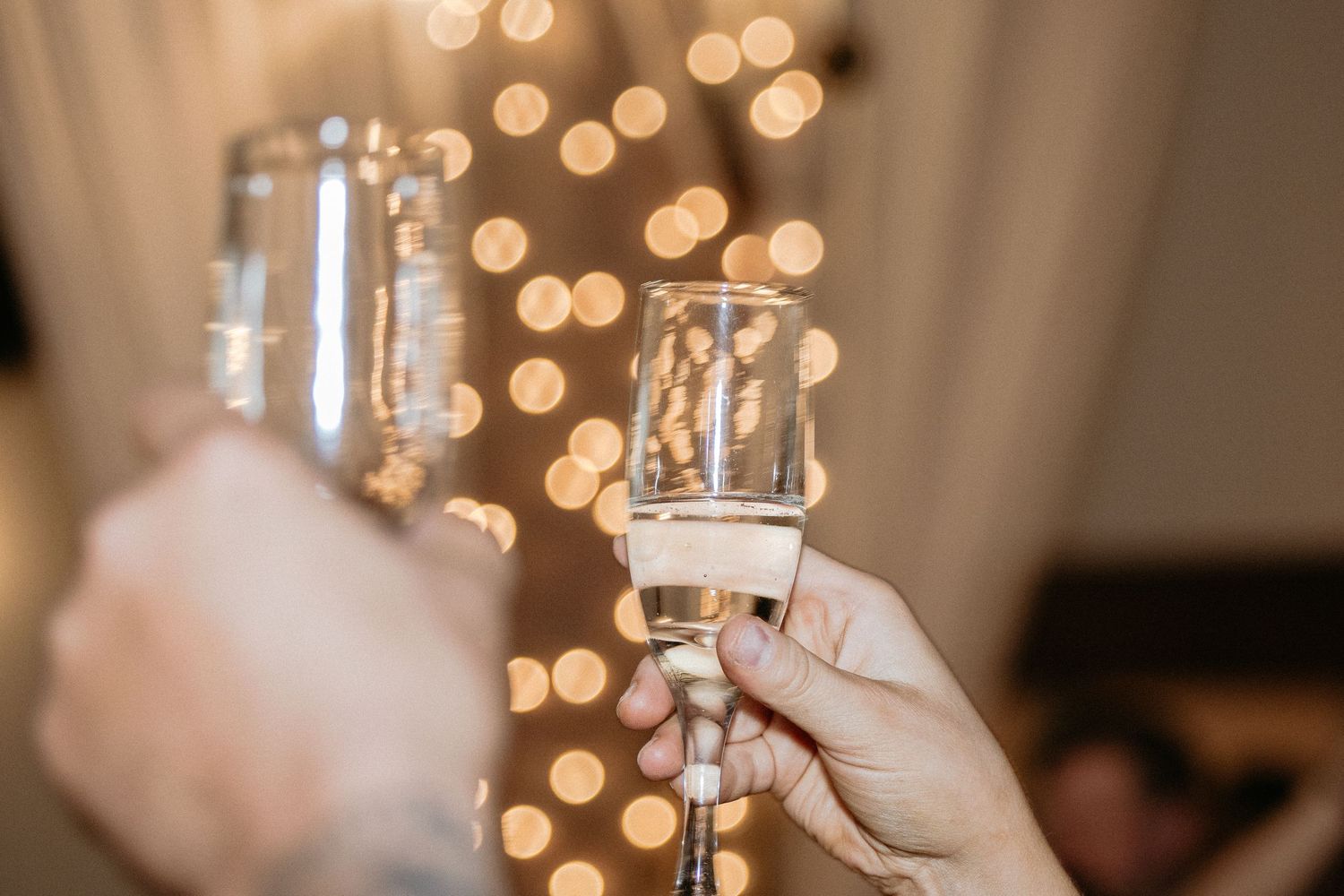 Hands hold champagne flutes for a wedding toast against twinkling bokeh lights.
