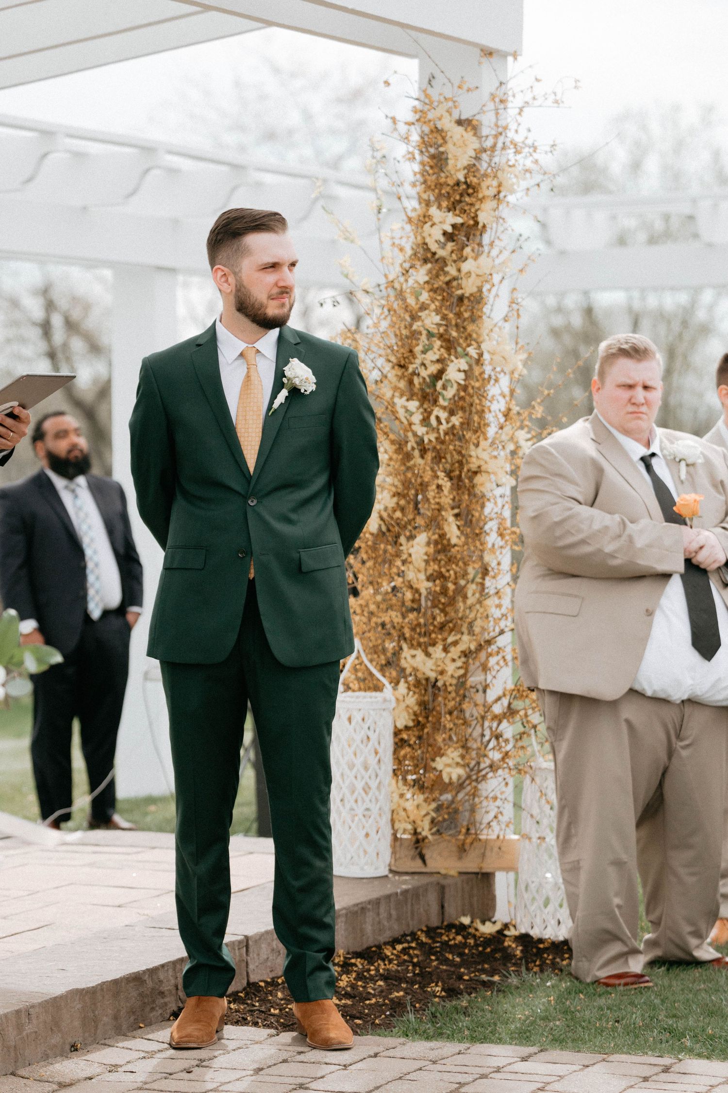 Groomsmen in dark green and tan suits stand together during an outdoor wedding ceremony.