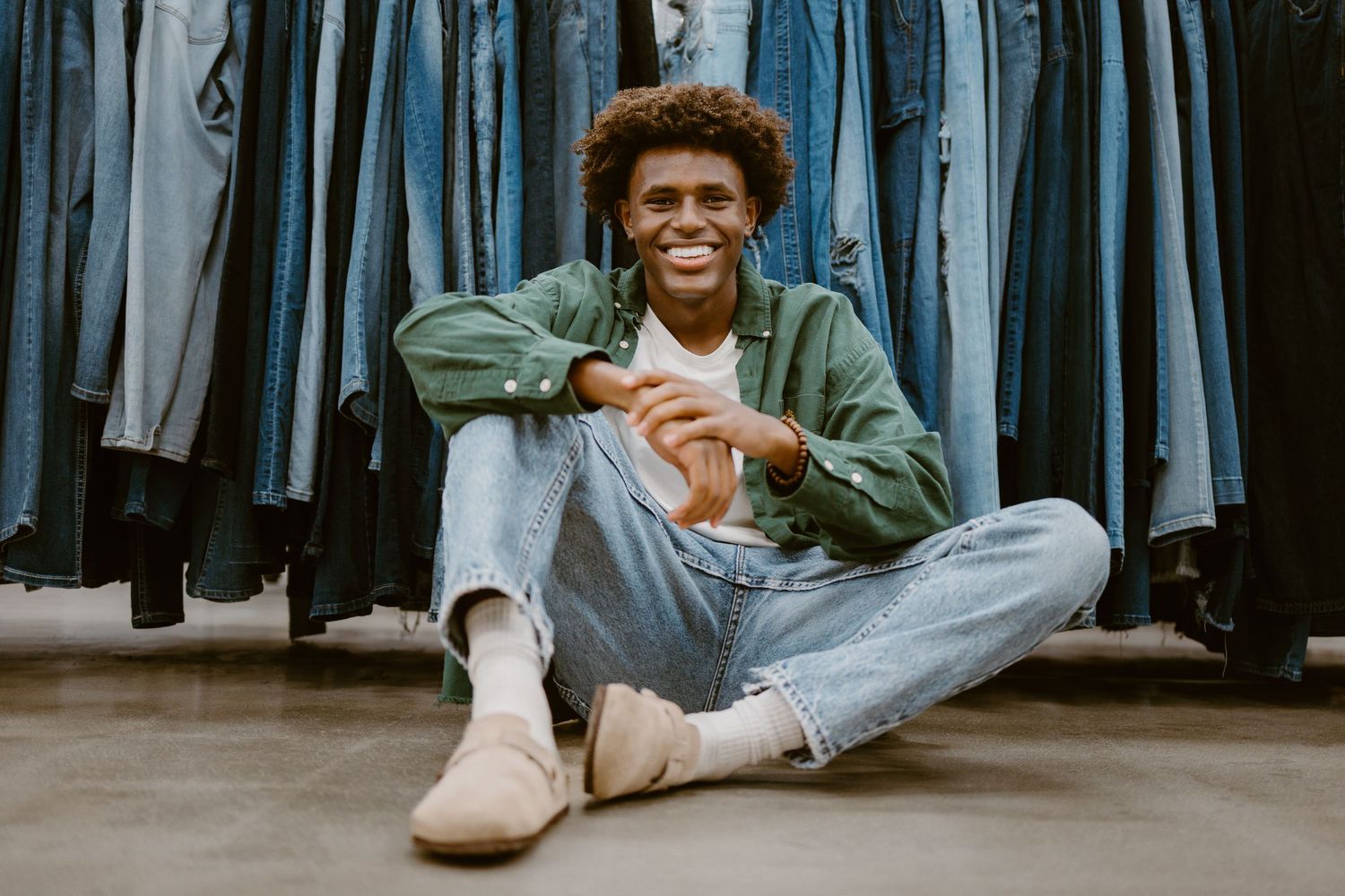 Person sitting on floor in shoe store surrounded by hanging clothing.