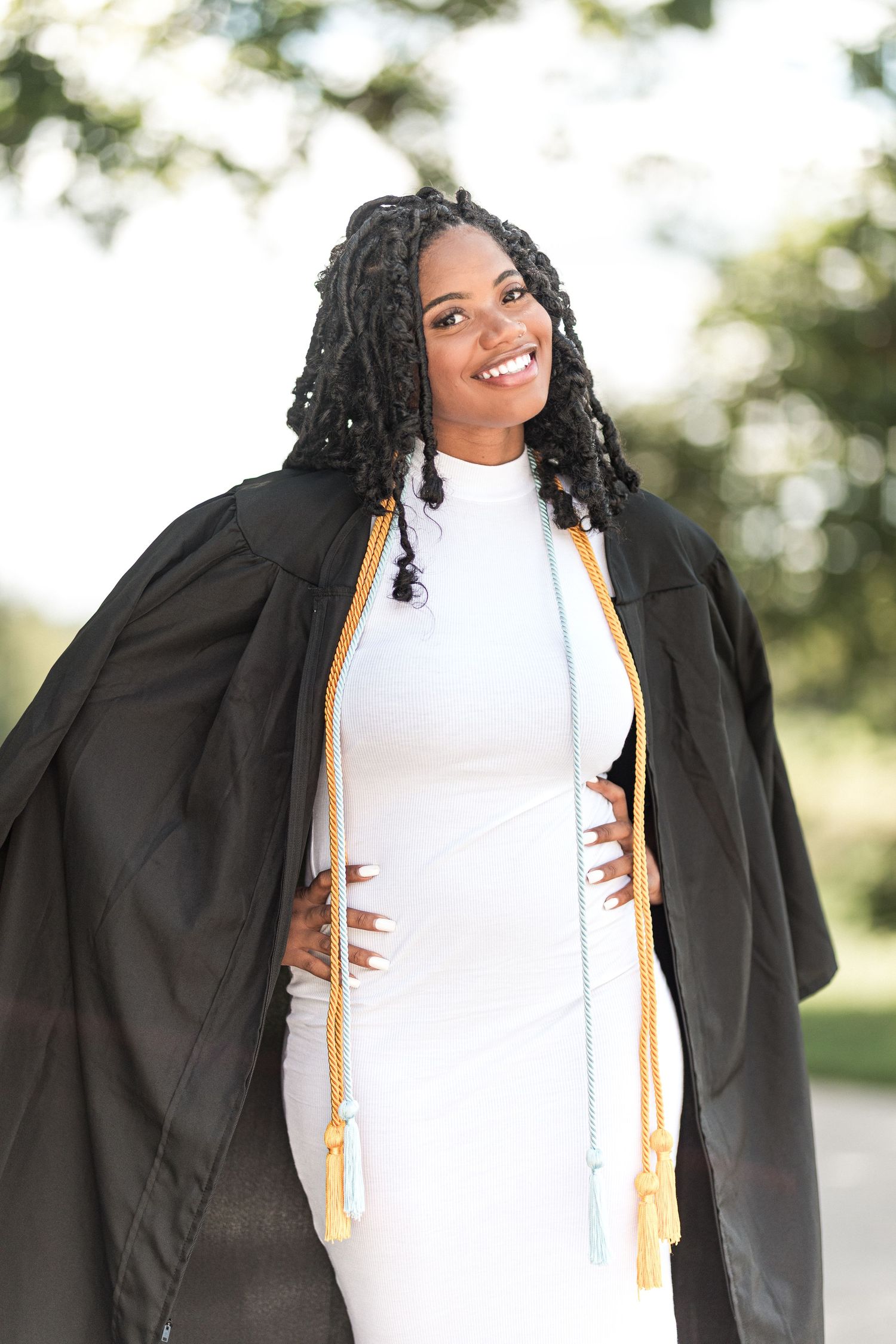 Graduate in black cap and gown with gold honors cords smiles joyfully while posing outdoors during graduation photo shoot.