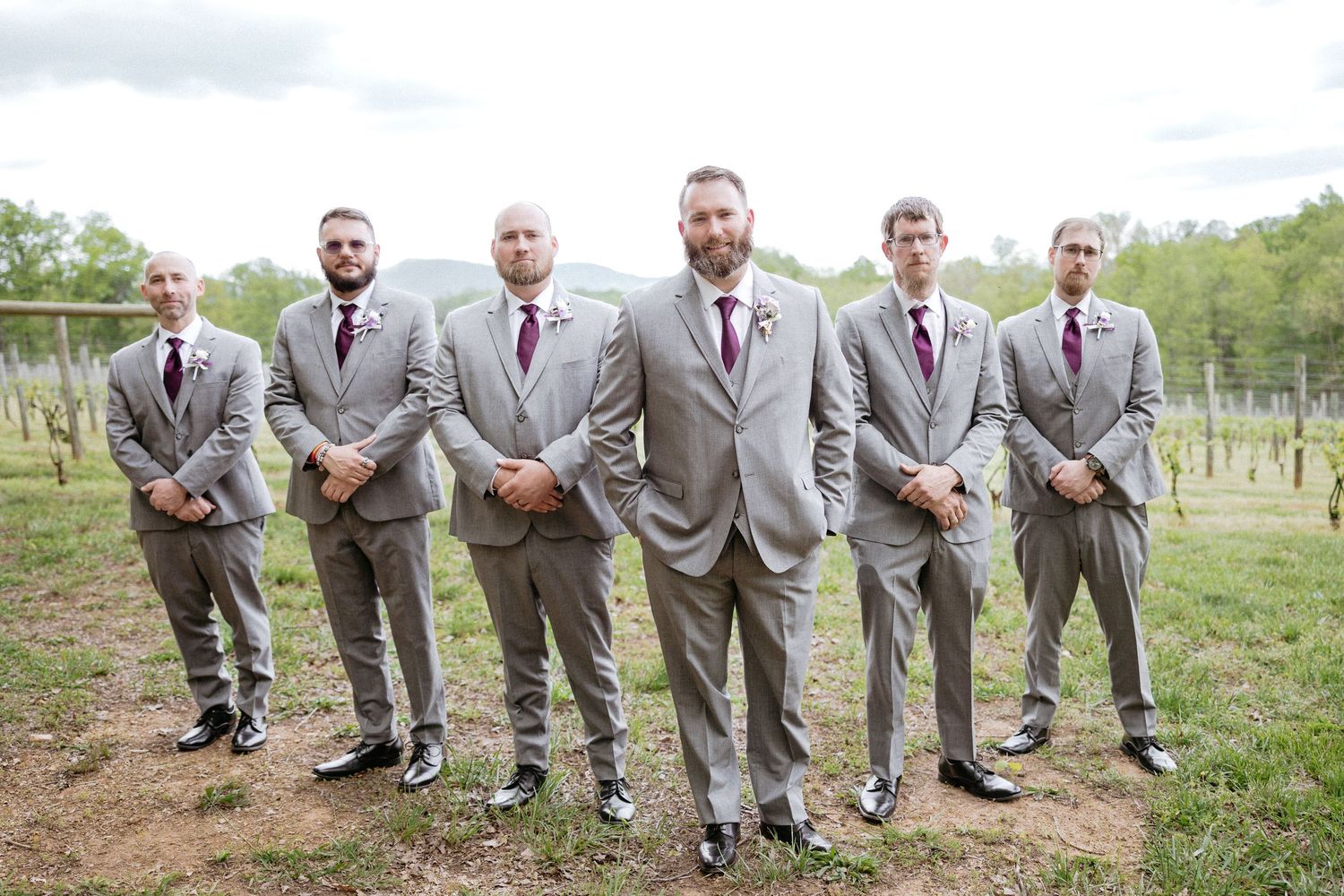 Groomsmen in matching gray suits show off their colorful socks while seated on grass outdoors.
