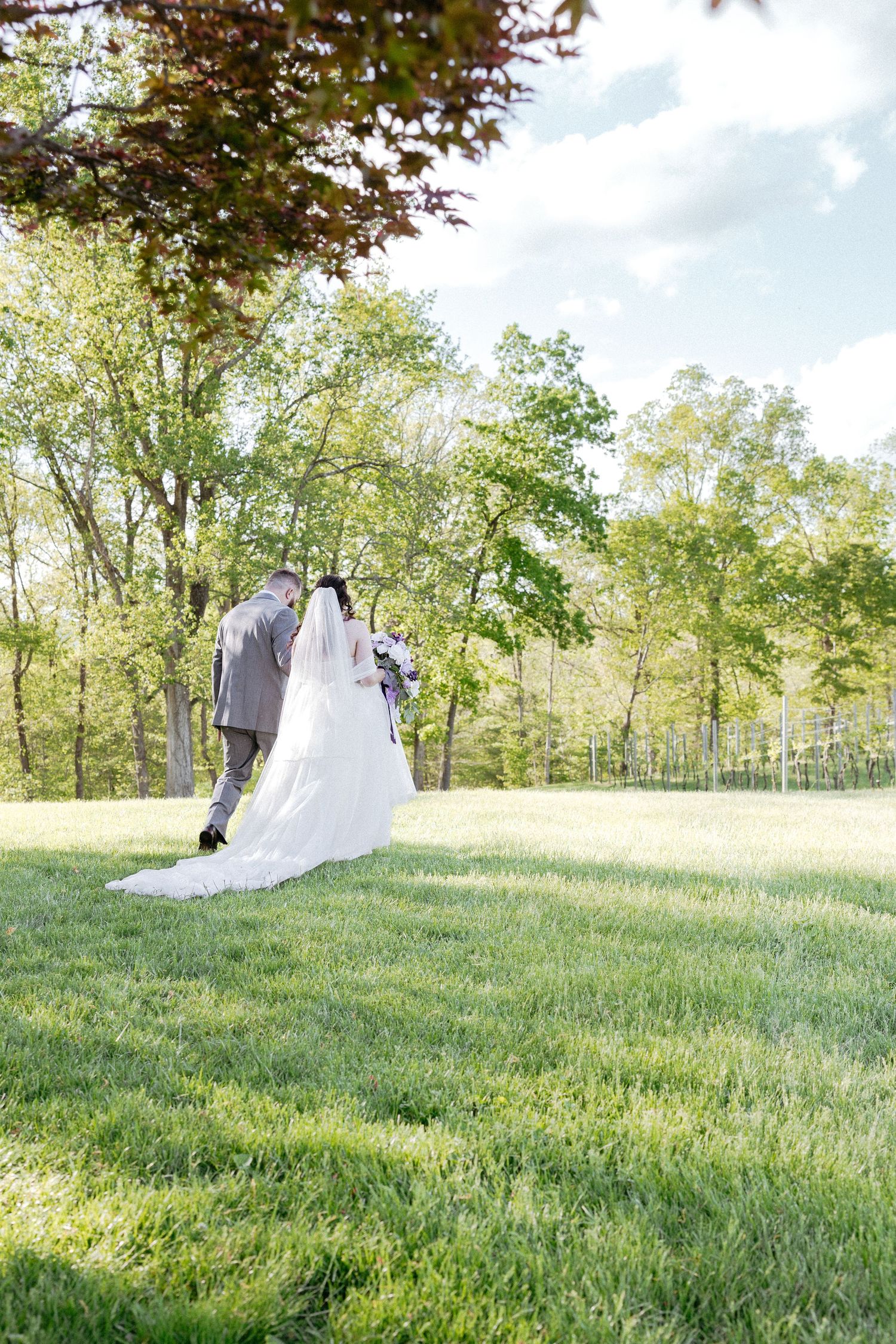 A flowing white wedding dress and veil billow elegantly across a green grassy hillside on a sunny day.