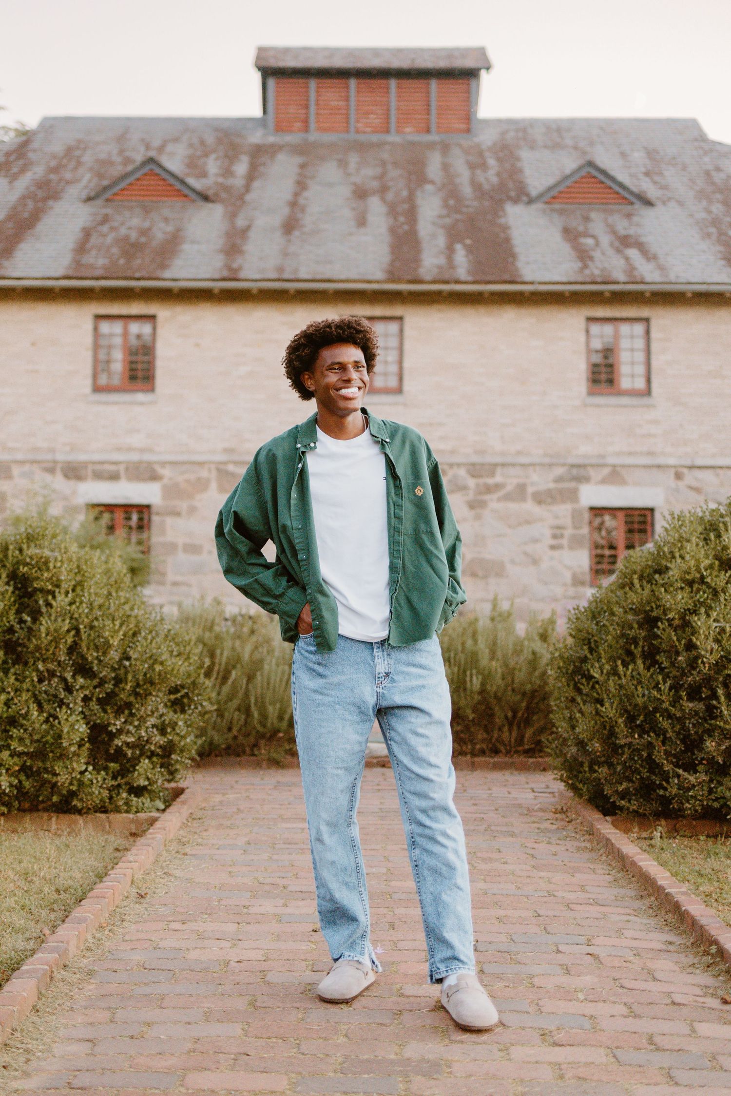Person in green jacket and light denim jeans poses playfully on stone walkway in front of historic stone building.