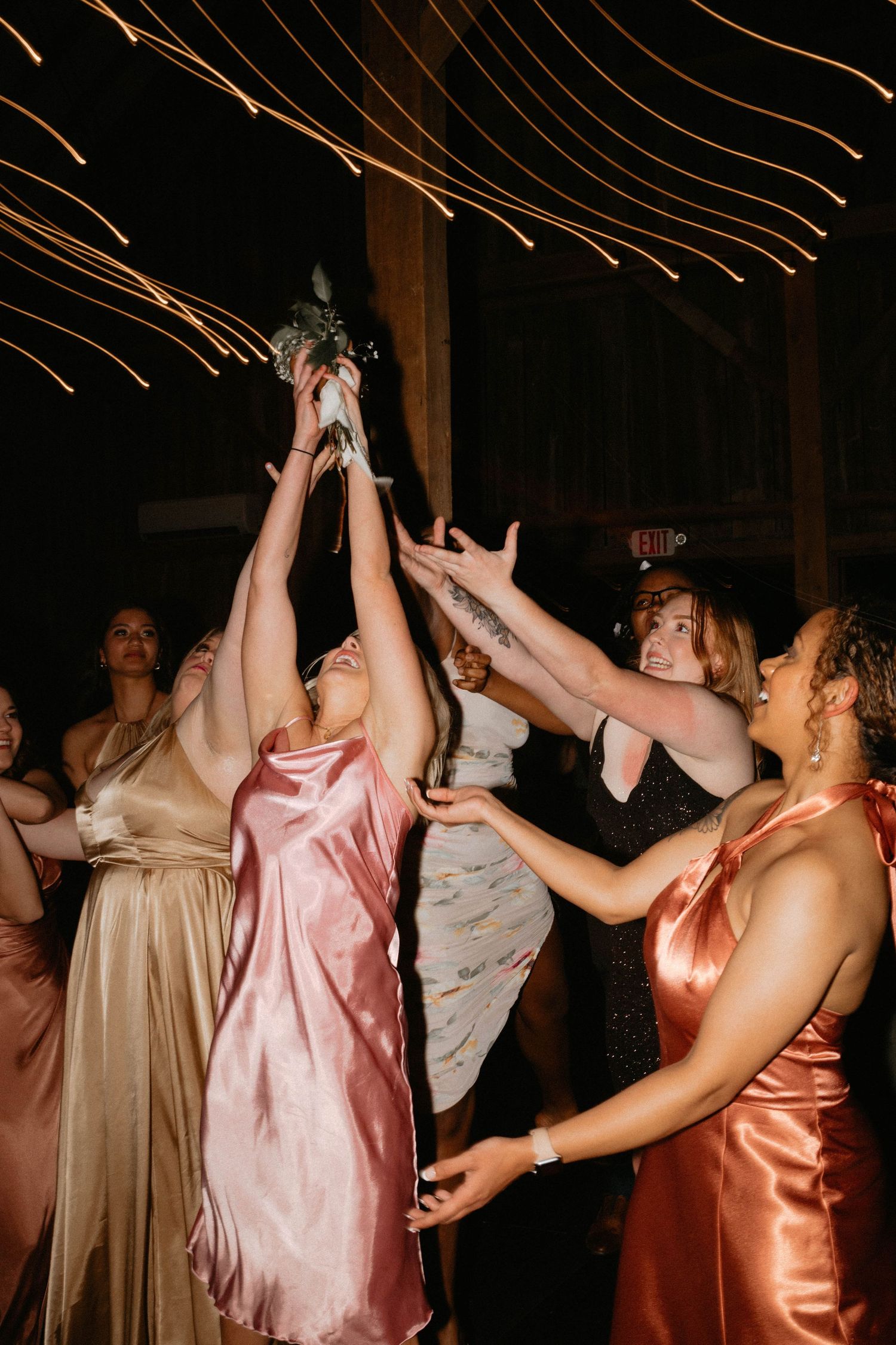 Wedding guests in satin dresses reaching up to catch bouquet during reception.