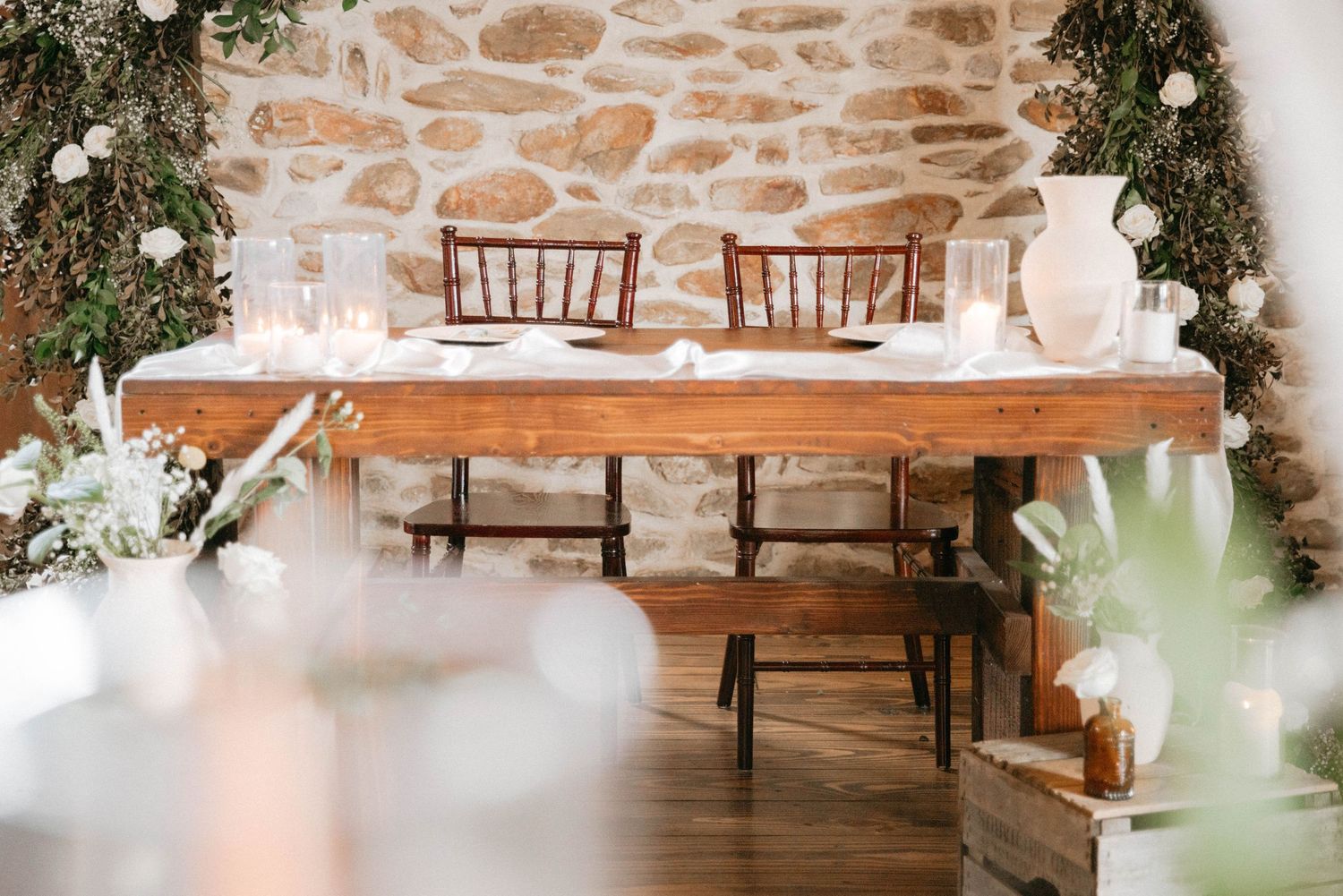 Elegant rustic reception space with wooden tables and stone walls decorated with greenery and lights.