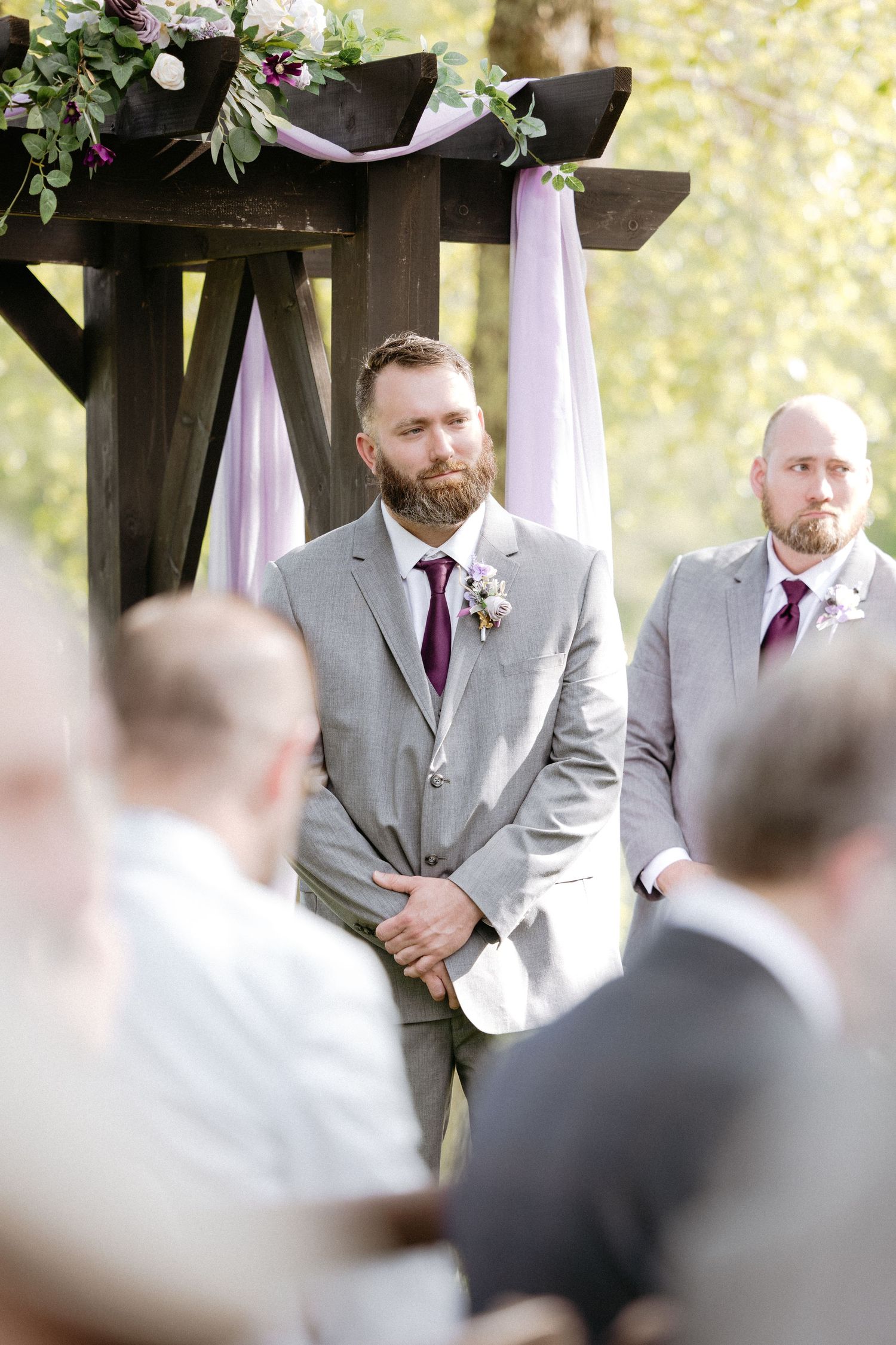 Groomsmen in light gray suits and burgundy ties stand in an outdoor wedding ceremony setting.